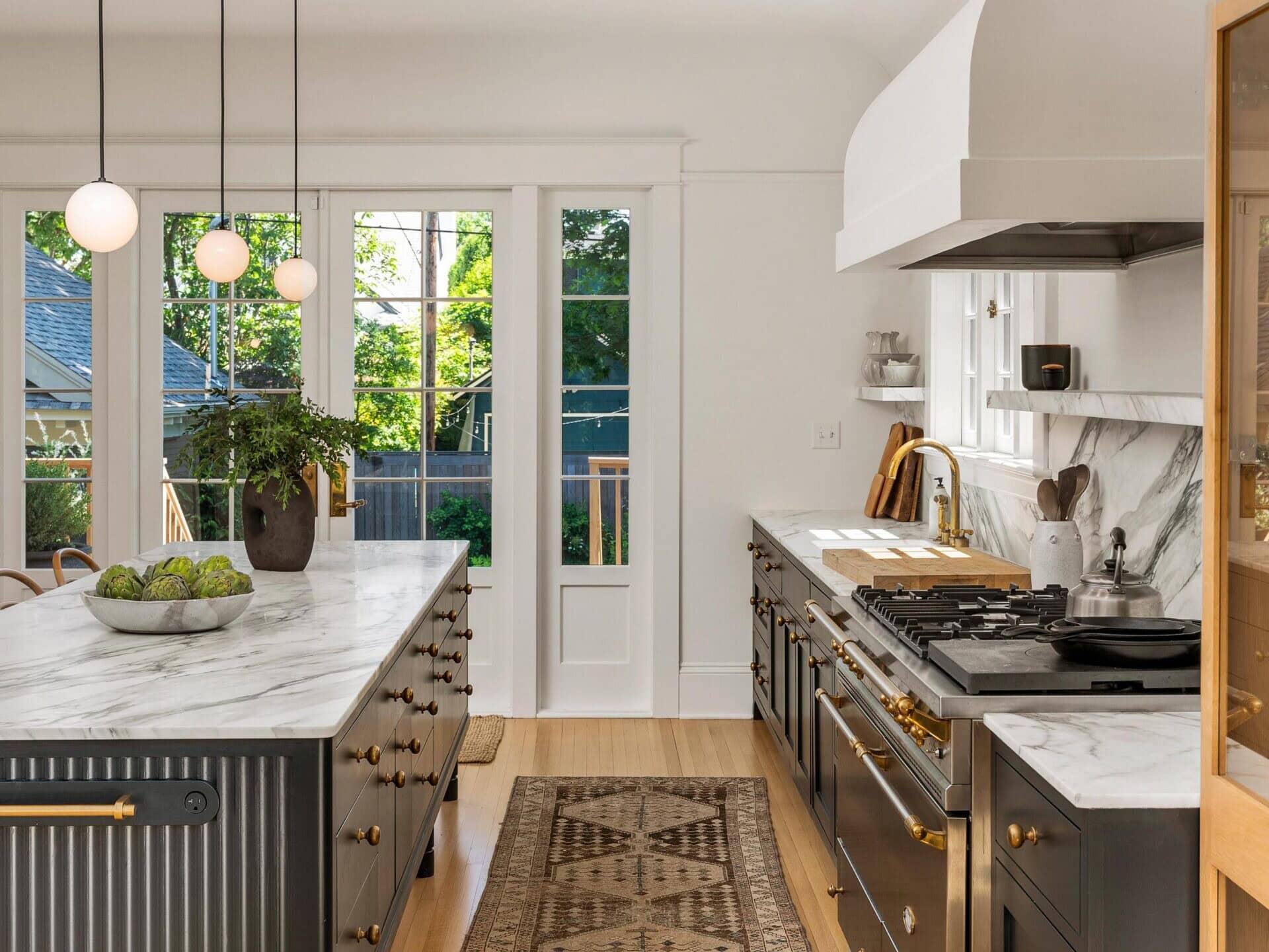 Bright, modern kitchen with black cabinets, marble countertops, a patterned rug, and a large island with pendant lights. Glass doors let in natural light and reveal greenery outside.