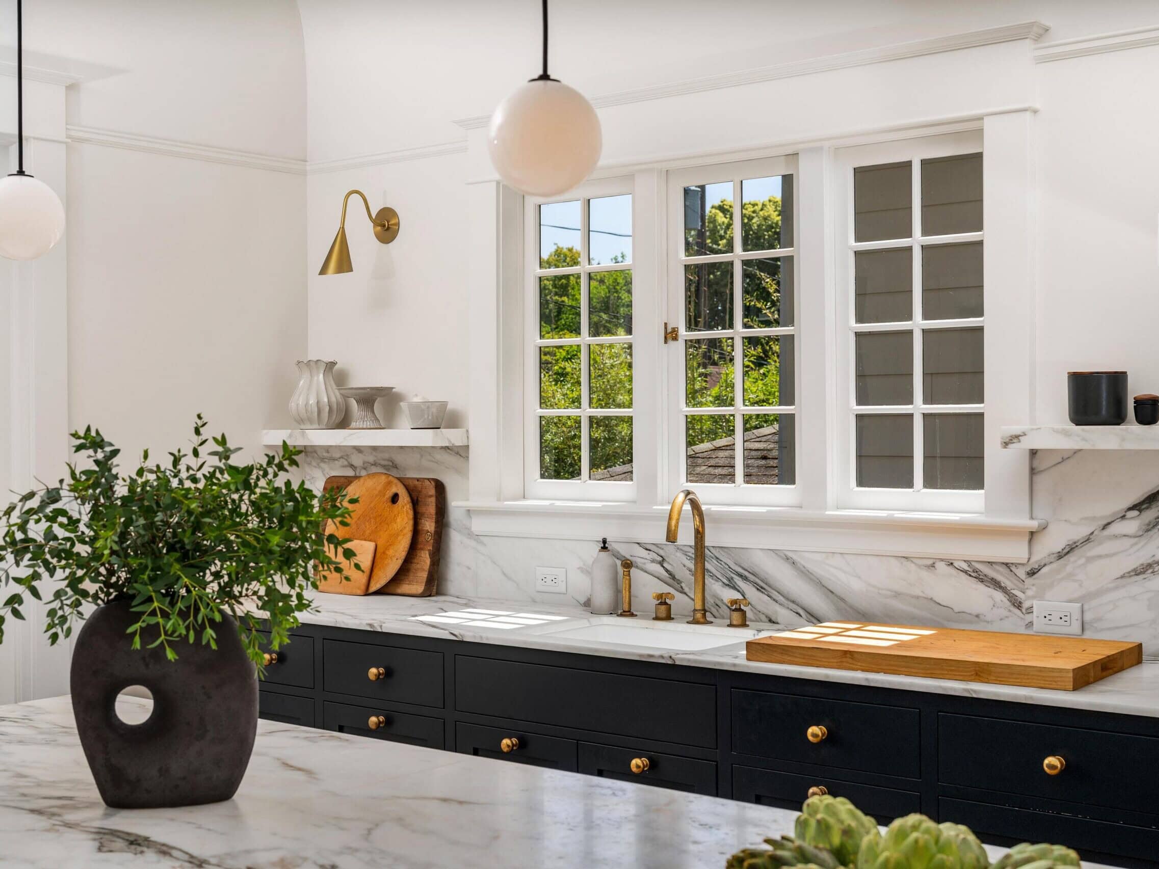 Bright, modern kitchen with white walls, marble countertops, black lower cabinets, gold faucet, and large windows. A potted plant sits on the island; cutting boards and dishes are visible near the sink.