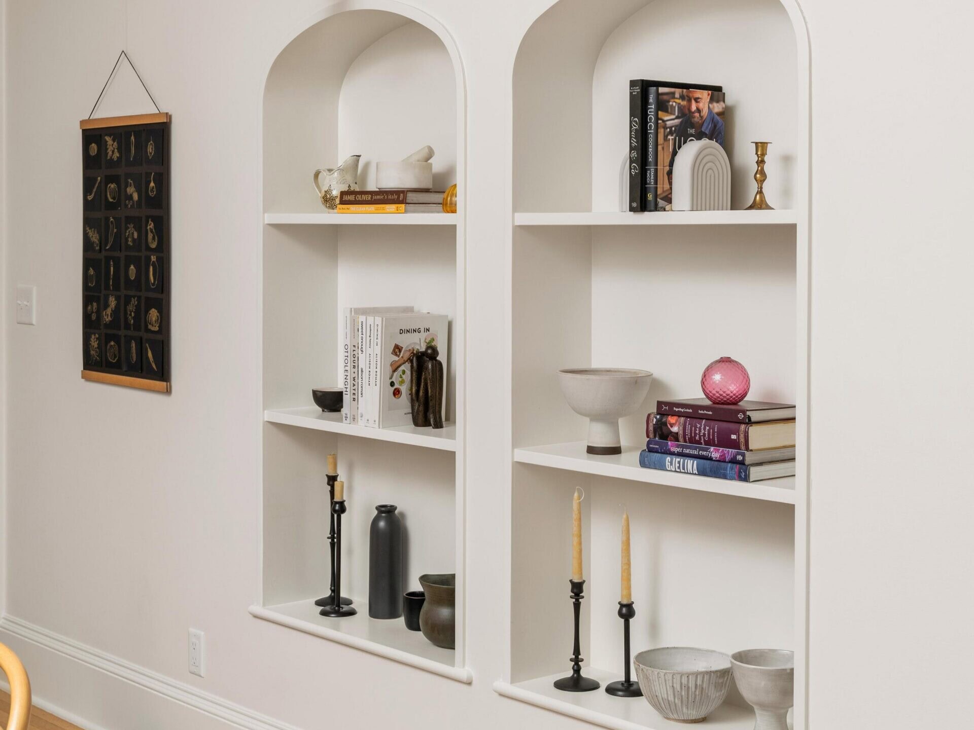 Two arched wall niches with shelves display books, candles, vases, and decorative objects. Above each niche is a gold wall sconce, and wooden chairs and a marble table are partially visible in the foreground.