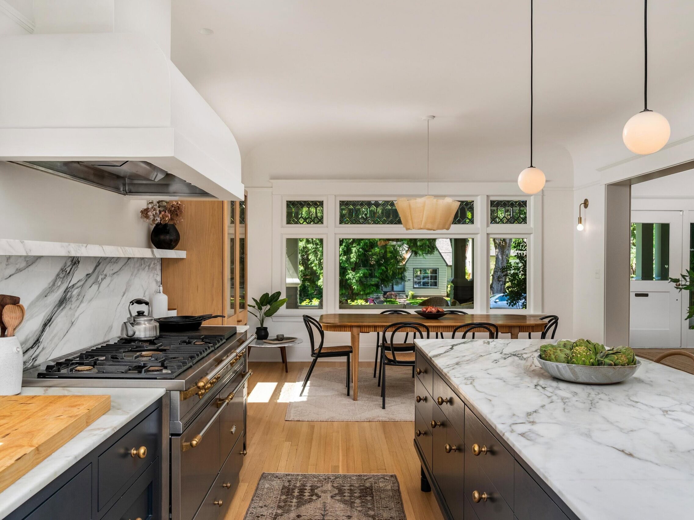 Bright, modern kitchen with marble countertops, black cabinets, and pendant lights. A stove with a kettle sits on the left, and a dining table with chairs is by large windows overlooking greenery.
