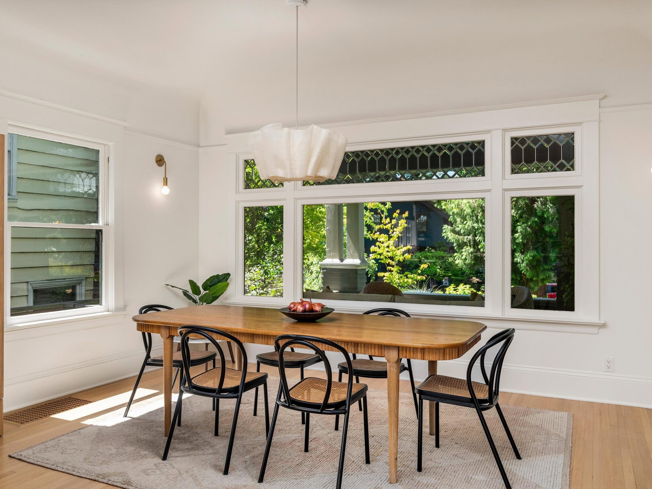 A bright dining room with a wooden table, six black chairs, a modern white pendant light, large windows showing greenery outside, and a light rug on a wood floor. A plant sits in the corner near the windows.