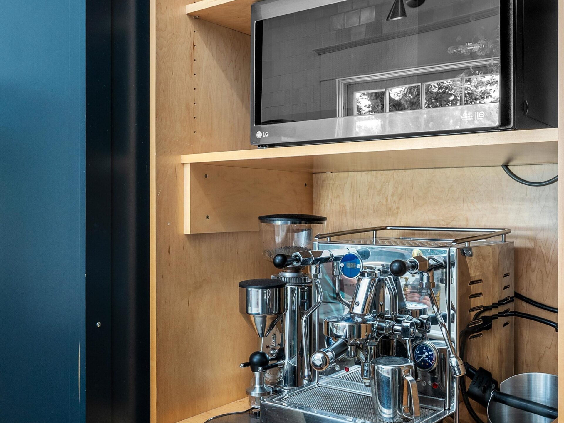 A shiny espresso machine sits on a wooden shelf below an LG microwave, with a jar of straws above and storage drawers below, all set inside a built-in dark blue kitchen cabinet.
