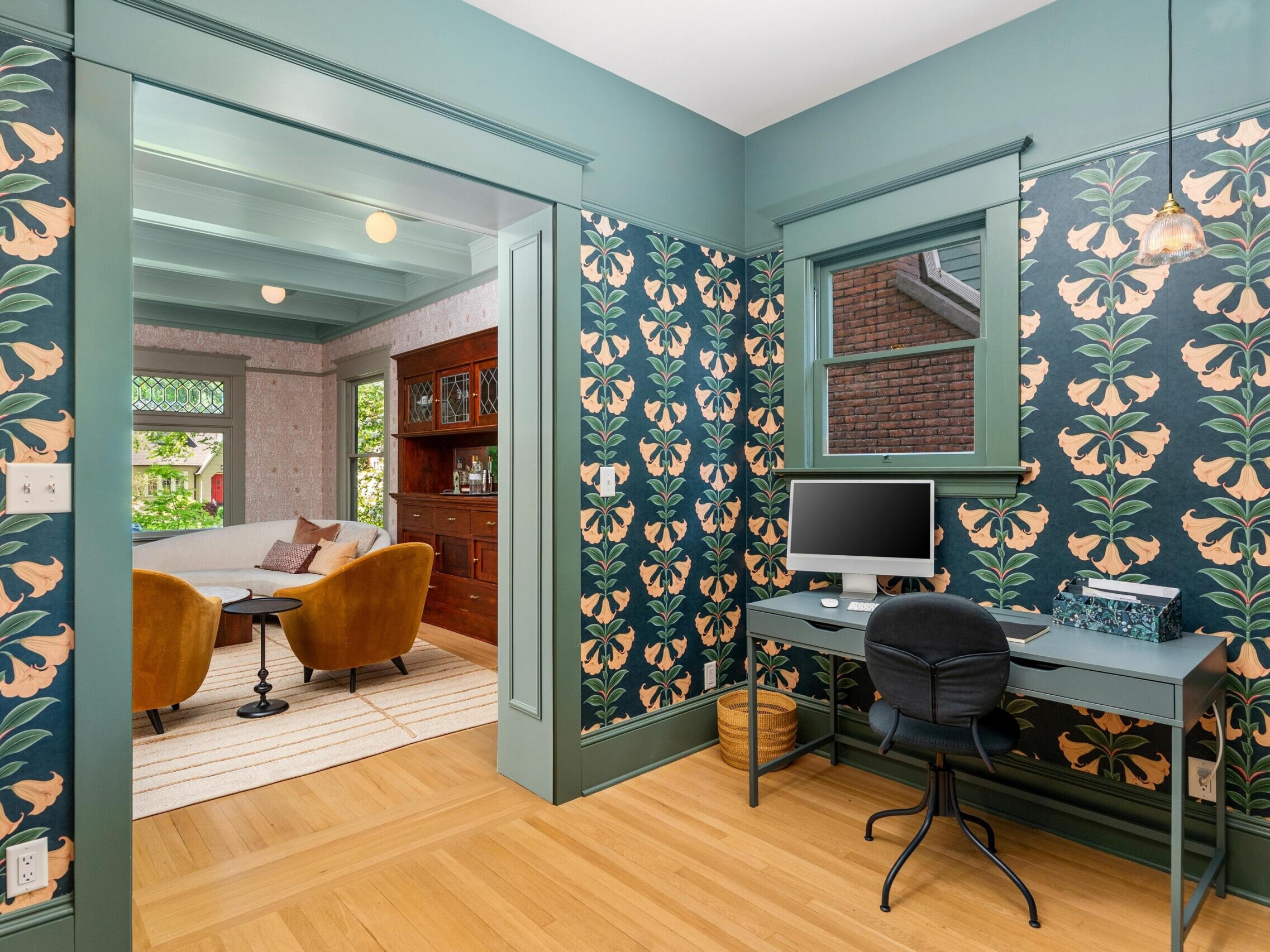 A home office with floral wallpaper, a desk with a computer, and a black chair. The room opens into a living space with two yellow armchairs, a brown couch, wooden shelves, and large windows letting in natural light.