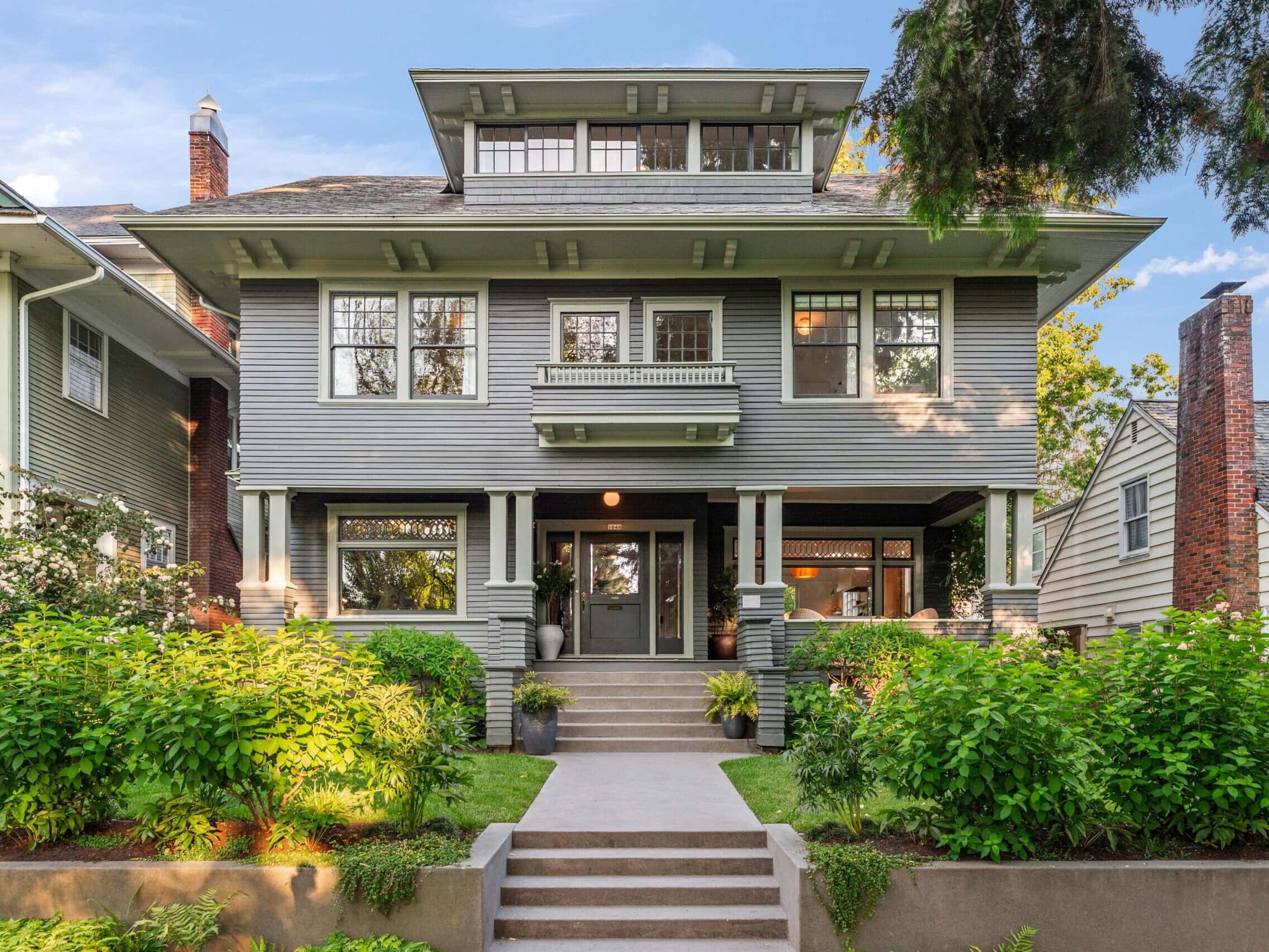 A large, two-story gray house with white trim, a covered front porch, and multiple windows, surrounded by lush green shrubs and plants, with a concrete walkway and steps leading to the entrance.