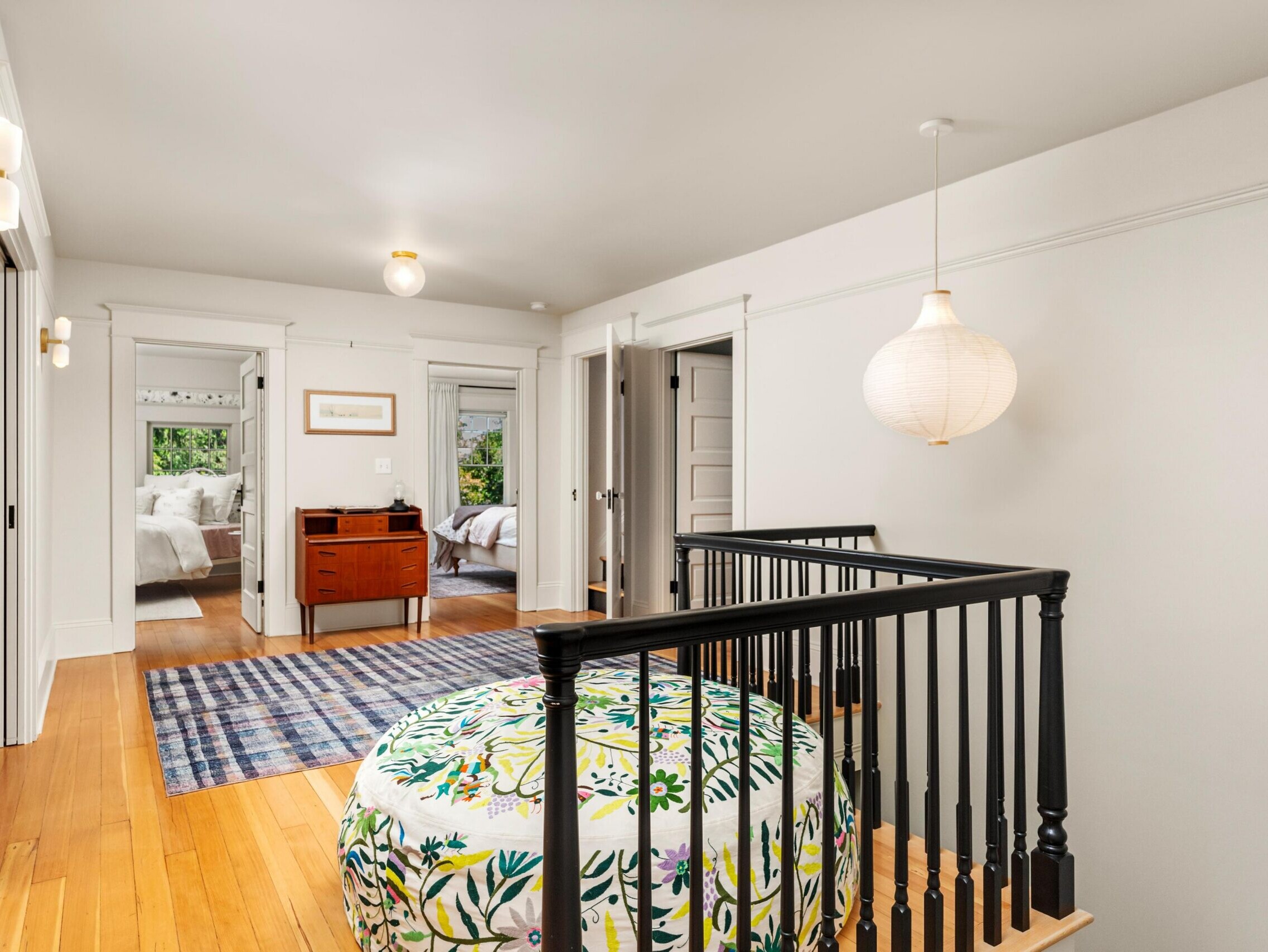 A bright upstairs hallway with light wood floors, white walls, and black railing around a staircase. A large round ottoman with a floral pattern sits on a rug. Two doorways lead to bedrooms, and a small wooden desk is against the far wall.