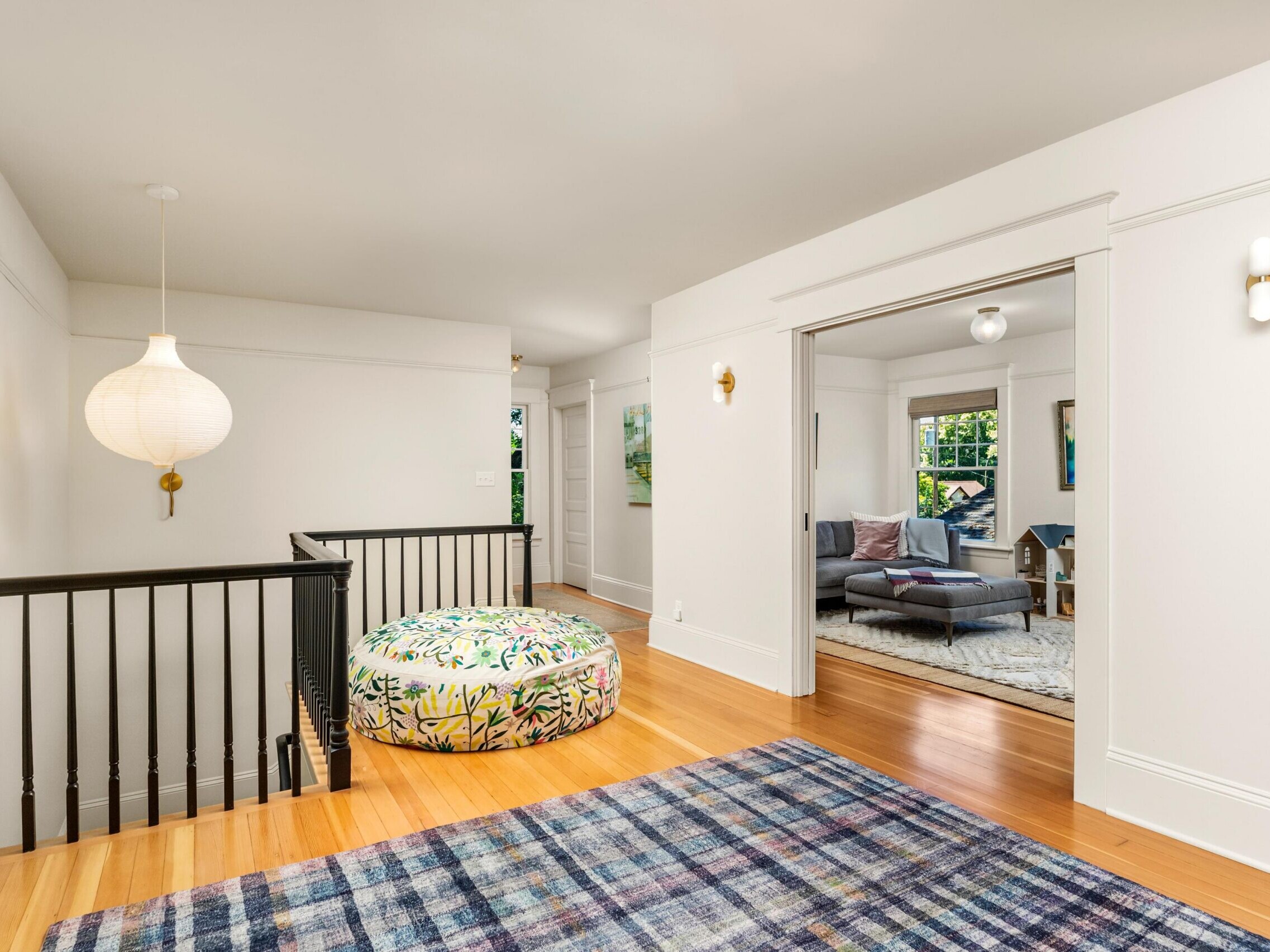 Bright upstairs landing with light wood floors, a patterned round ottoman, a blue plaid rug, white walls, black stair railing, a modern hanging lamp, and a view into a cozy living room with large windows.