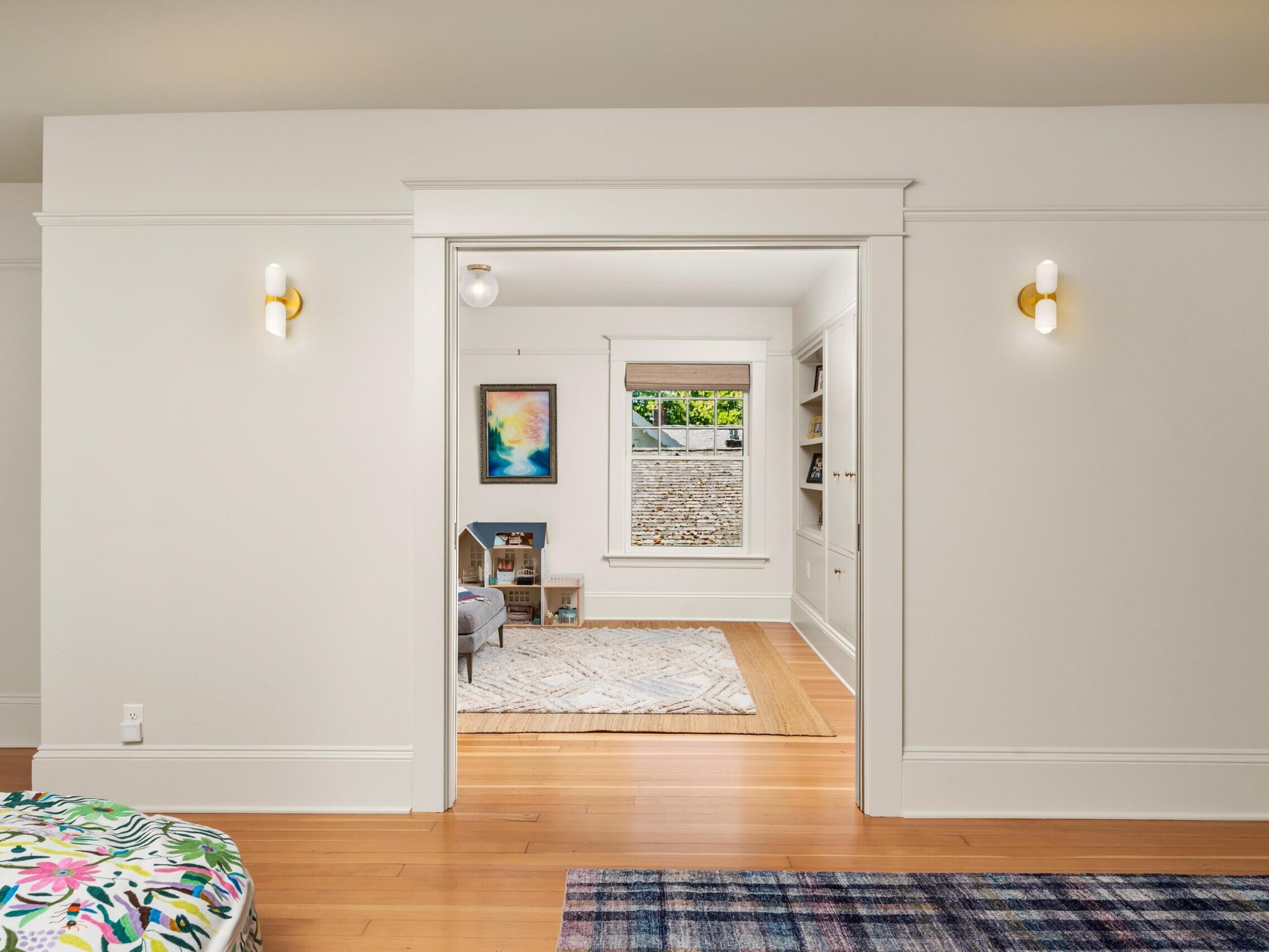 A bright room with white walls and trim opens to a cozy space with large windows, wooden floors, modern wall sconces, and colorful art and rugs. Built-in shelves and nature are visible through the windows.