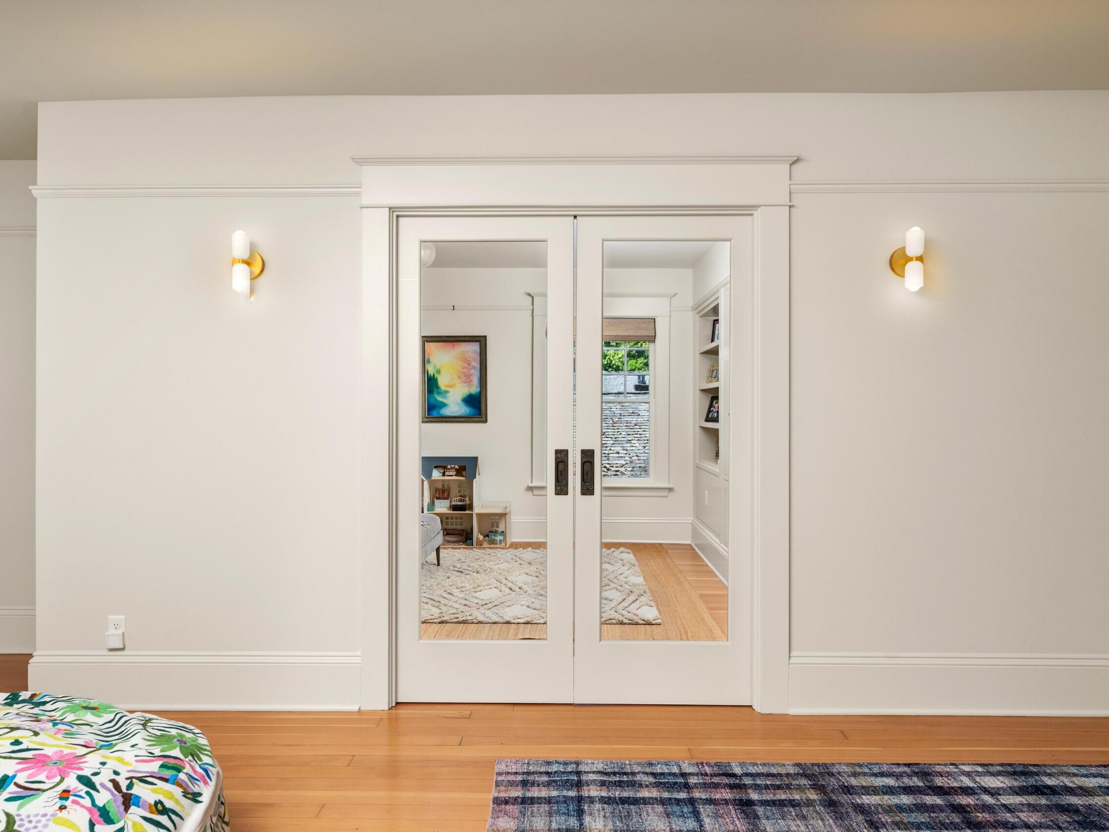 A bright room with wooden floors, white walls, and glass double doors leading to a cozy sitting area with art on the wall and bookshelves. Two modern wall sconces are on either side of the doors.