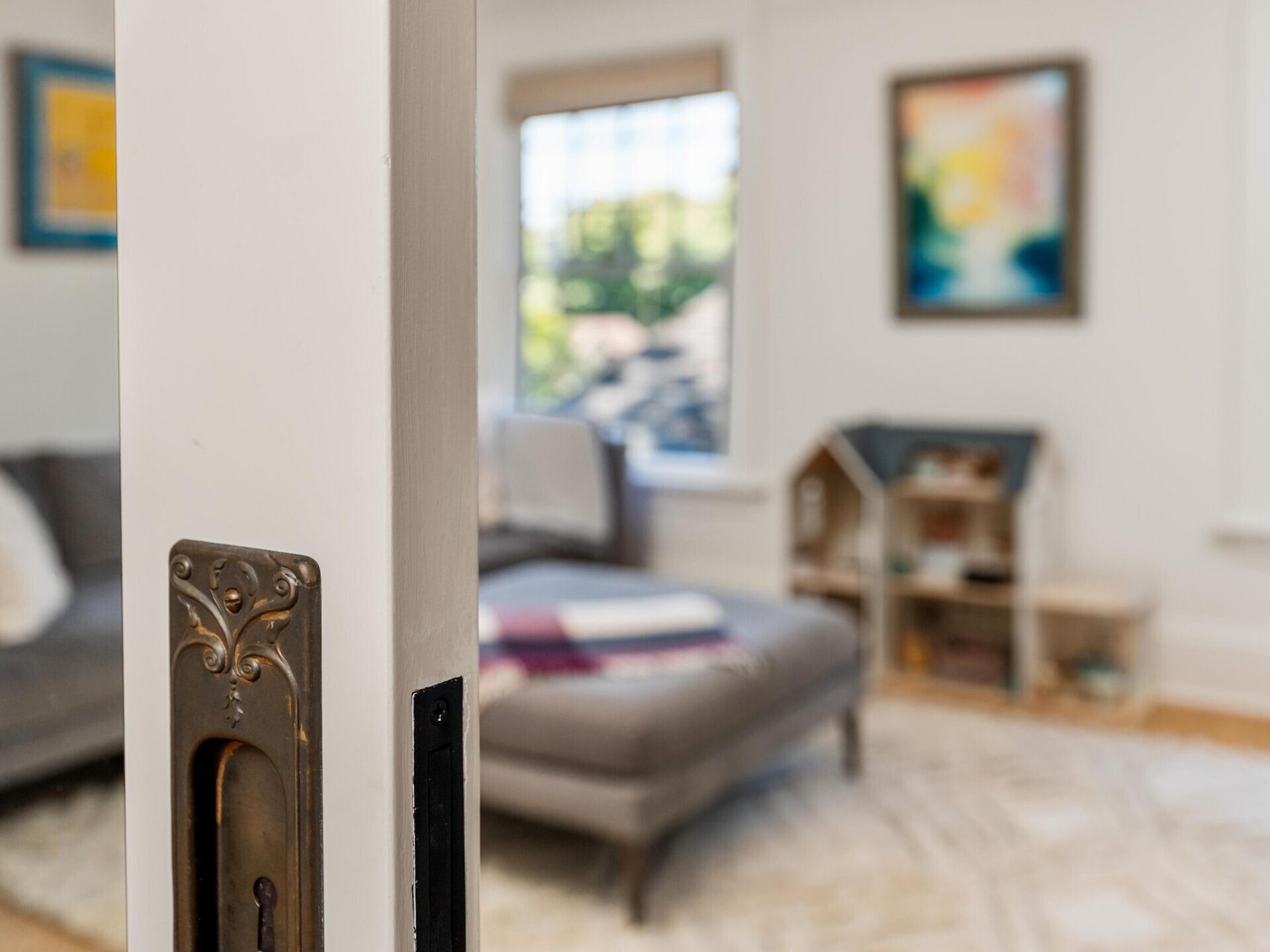 A close-up of an ornate brass door handle on a white door, with a blurred view of a living room featuring a sofa, rug, dollhouse, and colorful artwork in the background.