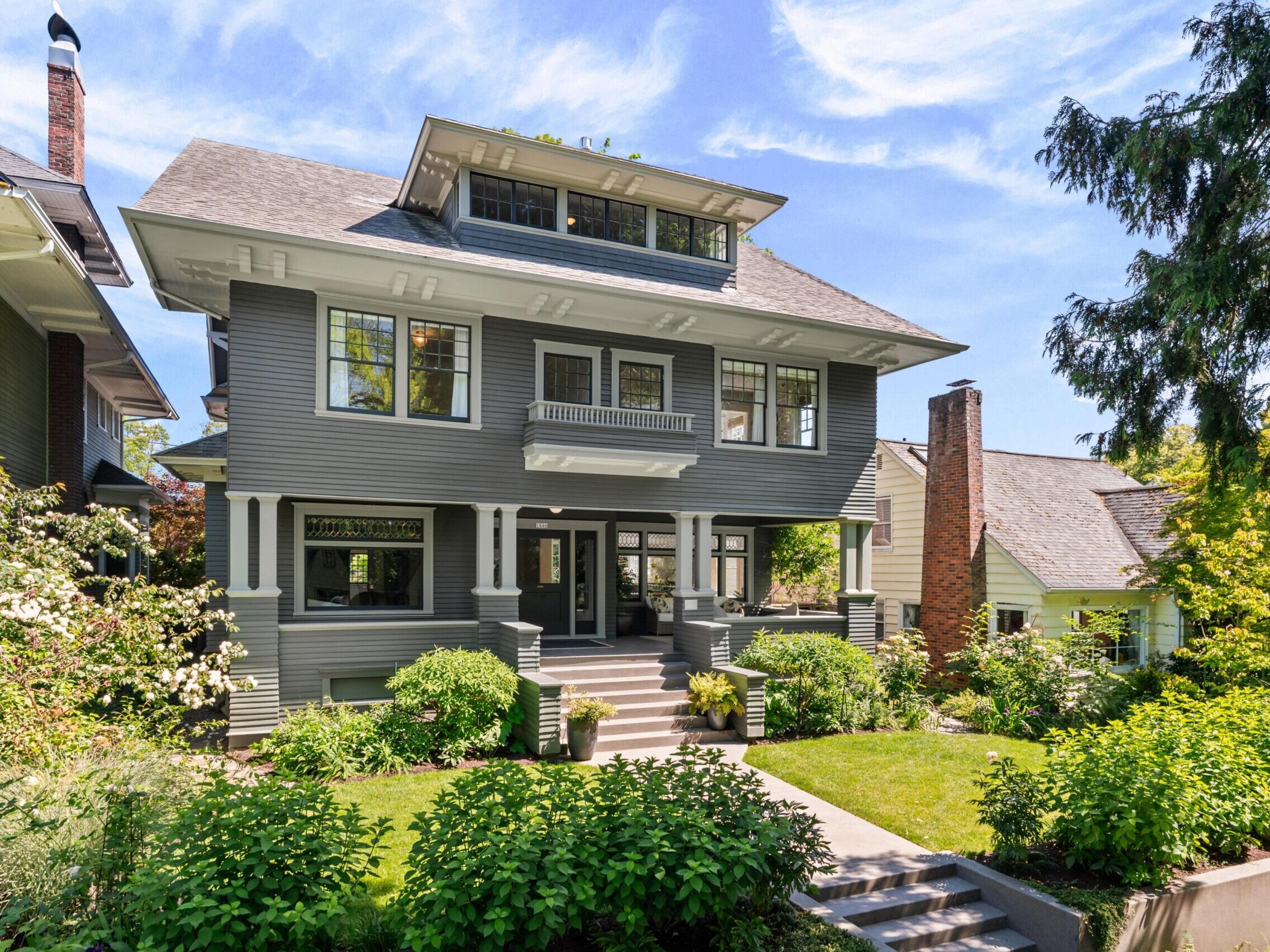 Gray Craftsman-style house with large windows, wide front porch, and neatly landscaped yard bordered by lush greenery, bushes, and mature trees on a sunny day. Stairs lead up to the welcoming entrance.