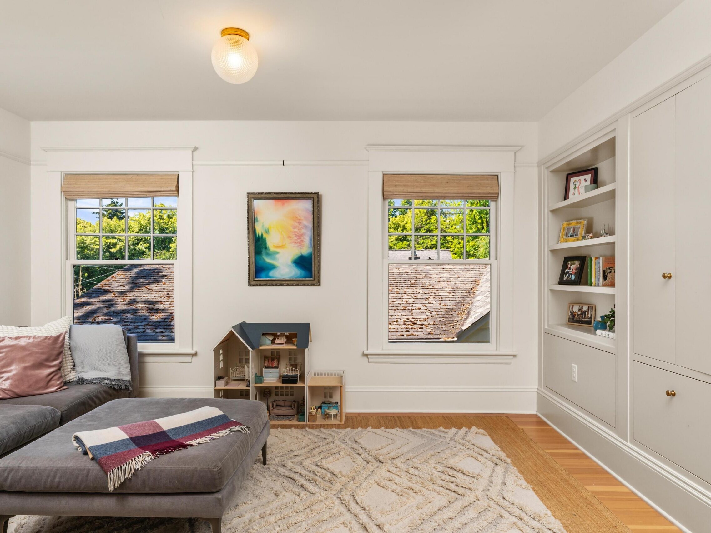 Bright living room with two windows, gray sectional sofa with pillows, ottoman with a blanket, built-in shelves with books and decorations, and a dollhouse beneath a colorful painting on the wall.