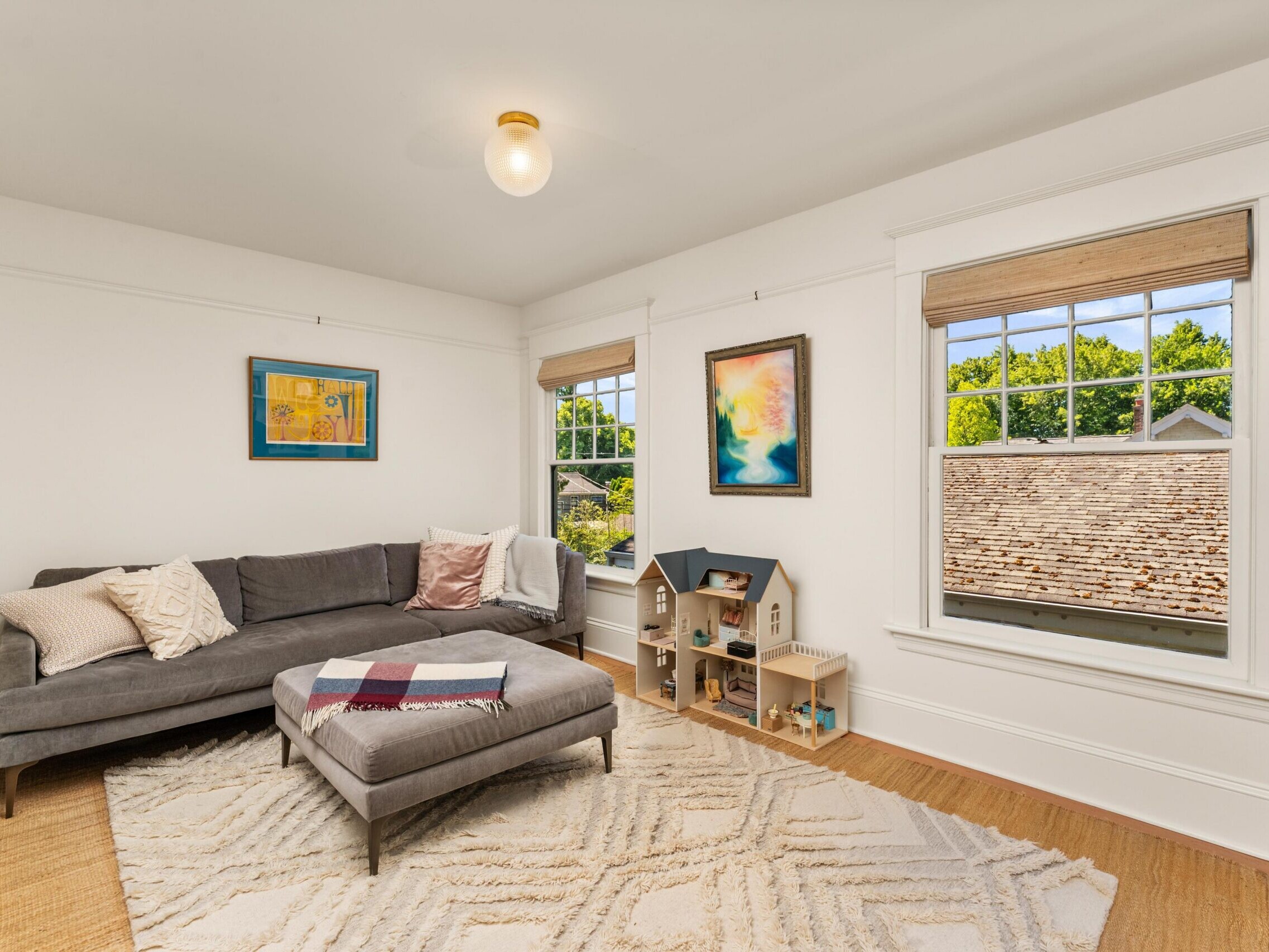 Bright living room with a gray sectional sofa, patterned ottoman, white rug, and a dollhouse by two large windows. Colorful artwork hangs on the white walls, and sunlight fills the space.