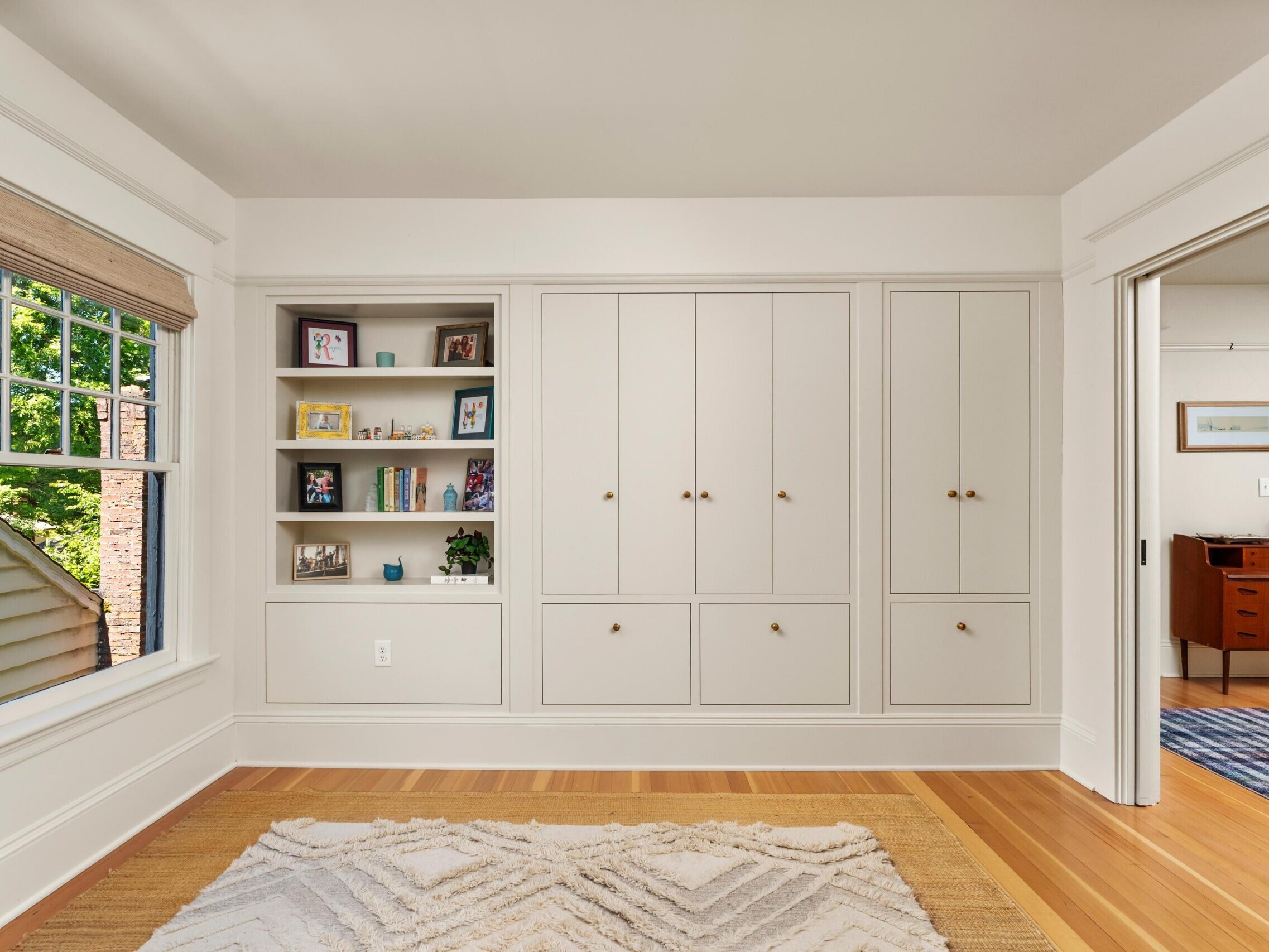 A bright room with a window, built-in shelves displaying books and decor, and large cabinets with gold knobs. A geometric-patterned rug covers the wooden floor, and sliding doors lead to another room.
