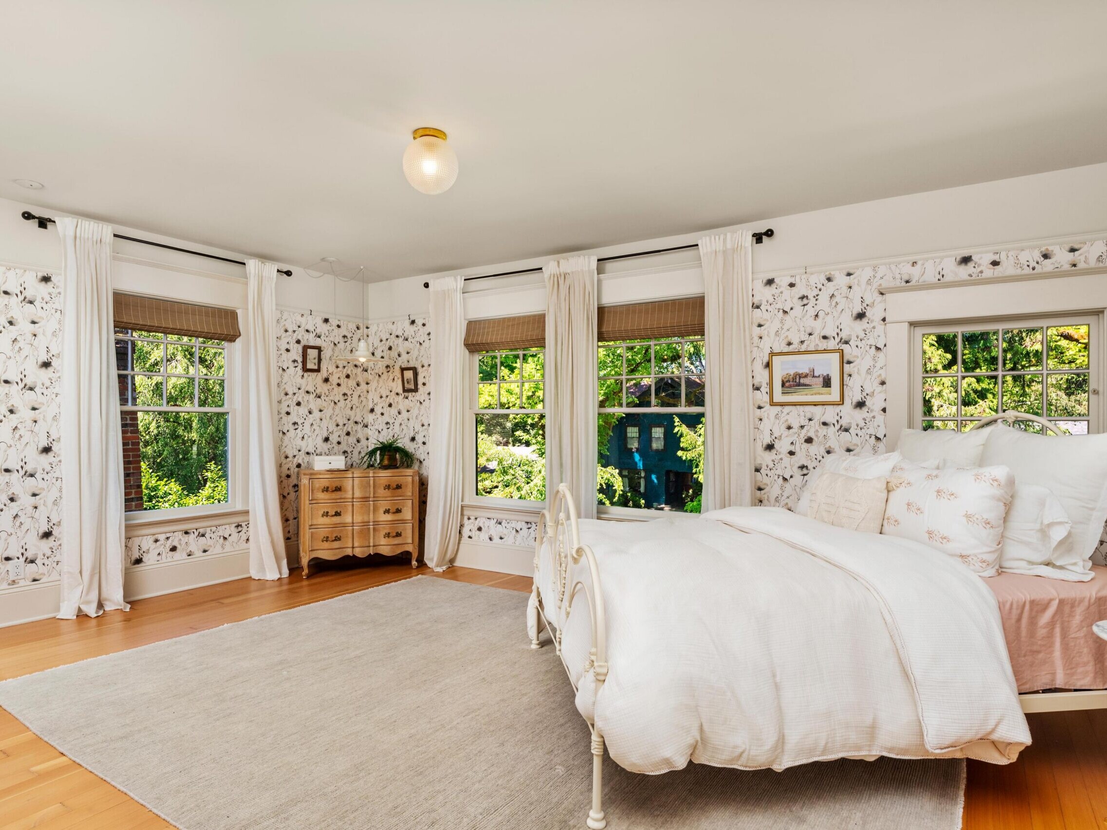Bright bedroom with floral wallpaper, three large windows with white curtains, a white metal bed with light bedding, a wooden dresser in the corner, light wood floors, and a neutral area rug. Lush greenery visible outside.