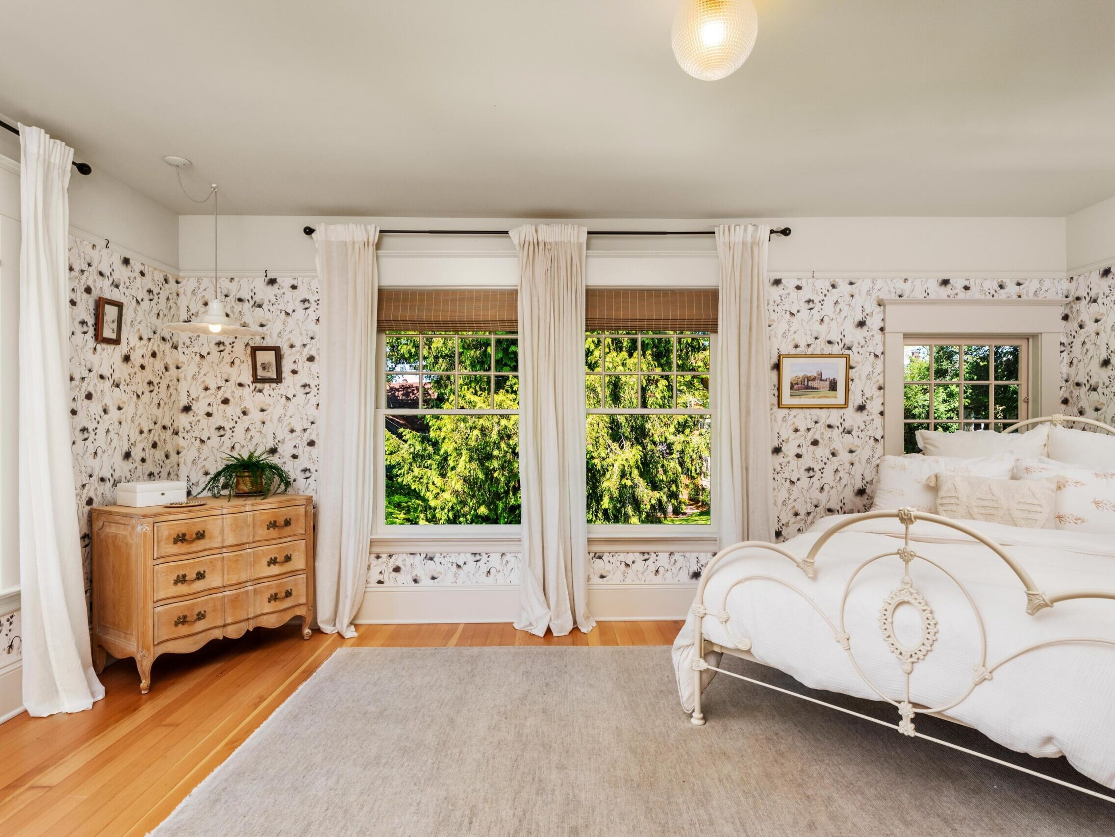 Bright bedroom with large windows, floral wallpaper, a vintage metal bed with white bedding, light curtains, wooden floors, a beige rug, and a wooden dresser with a plant. Lush greenery visible outside.