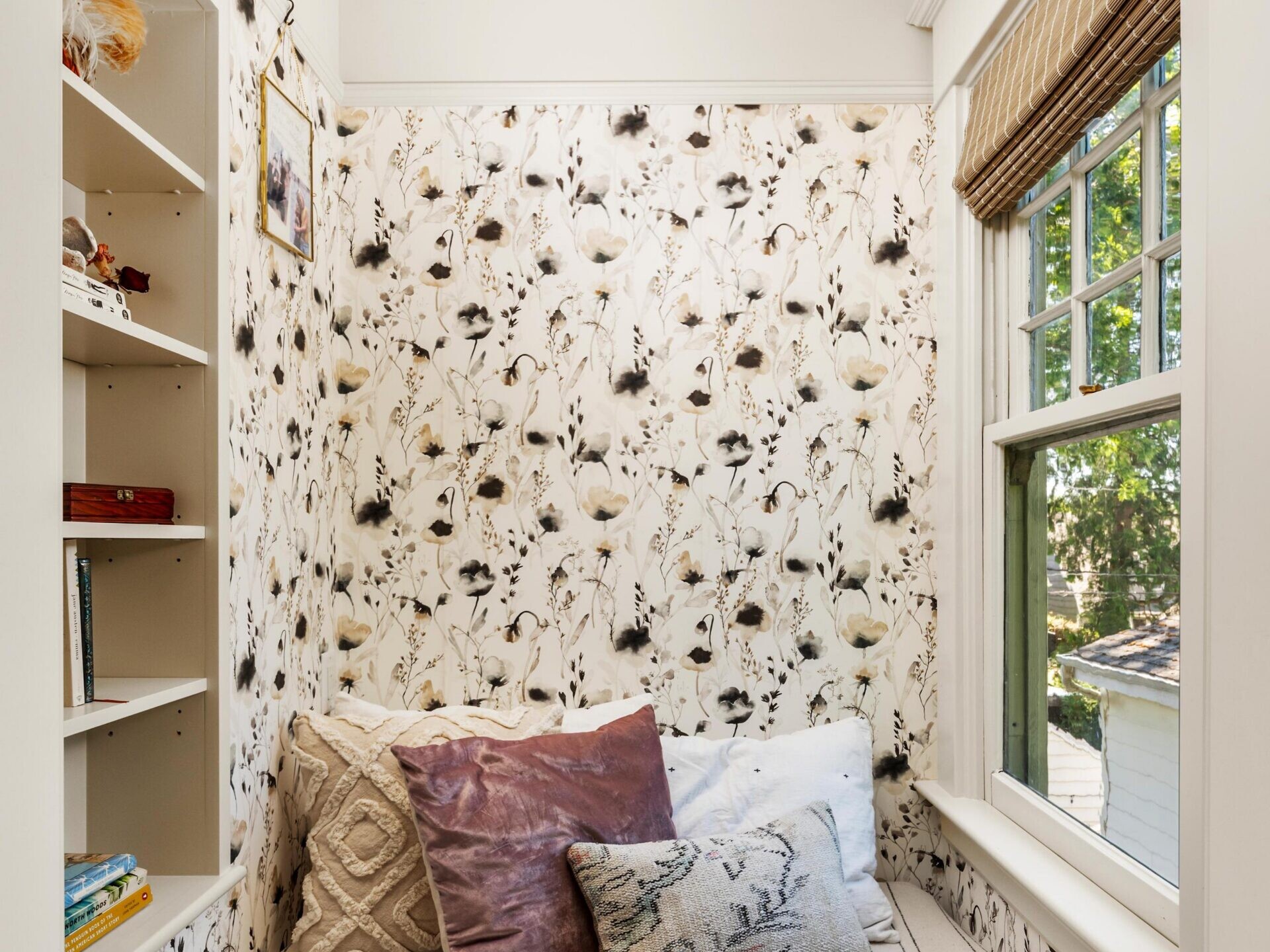 A cozy reading nook with patterned wallpaper, built-in shelves holding books and decor, large pillows on a striped cushion, and a window with natural light and an outdoor view.