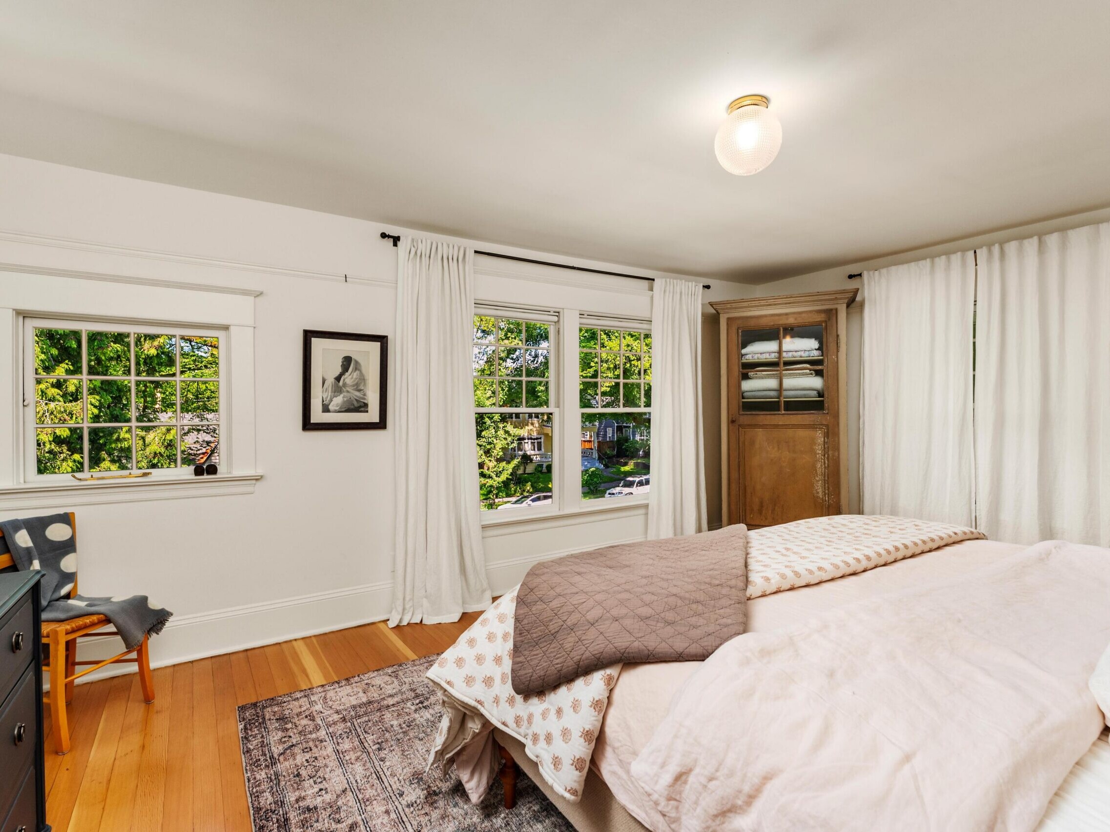 Bright bedroom with large windows, white walls, sheer curtains, a wooden bed with layered bedding, a patterned rug, a black dresser, a wooden wardrobe, a framed photo, and a chair by the window.