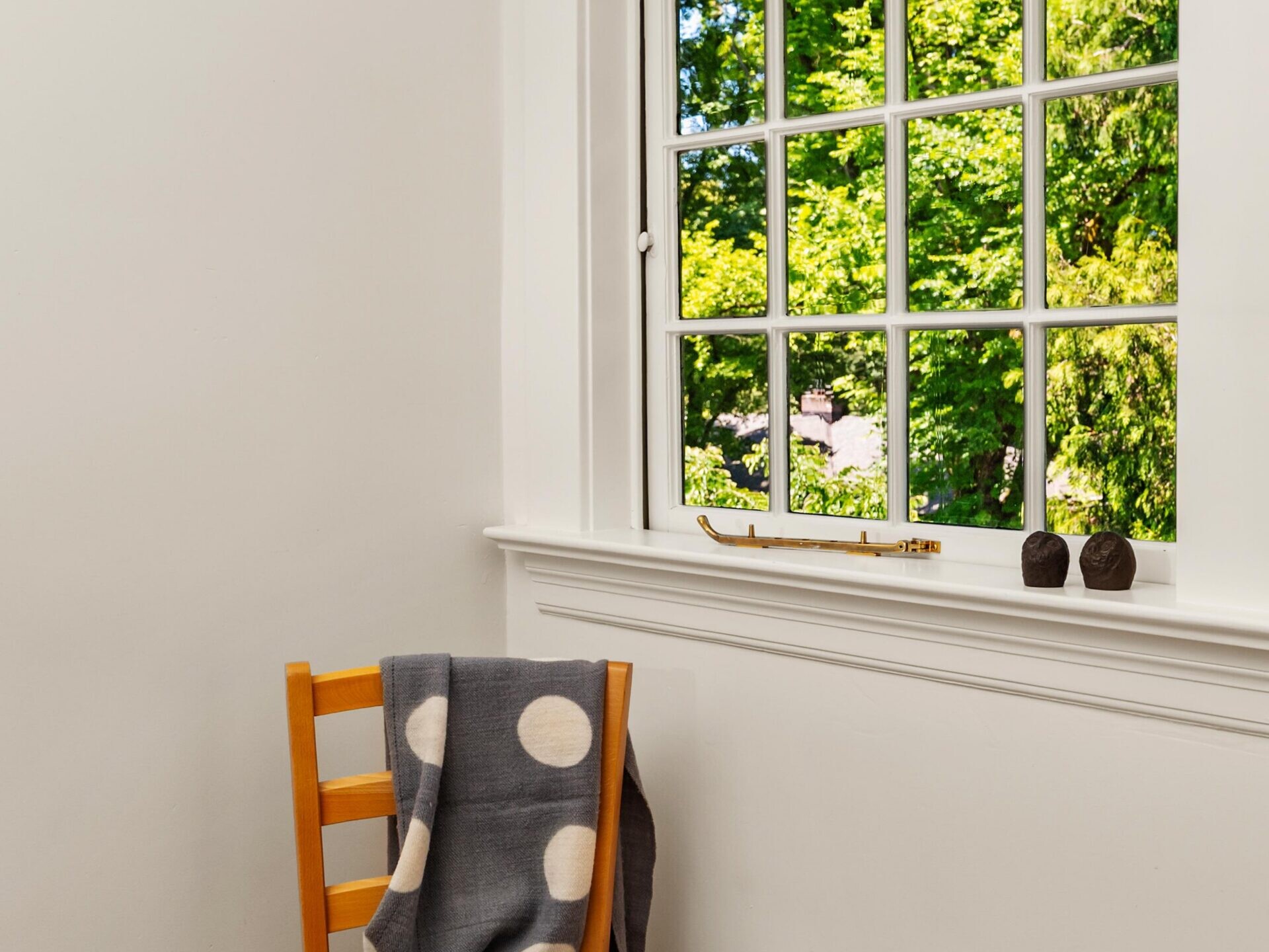 A wooden chair with a gray polka dot blanket draped over it sits by a window, which reveals lush green trees outside. The room has white walls and simple decor.