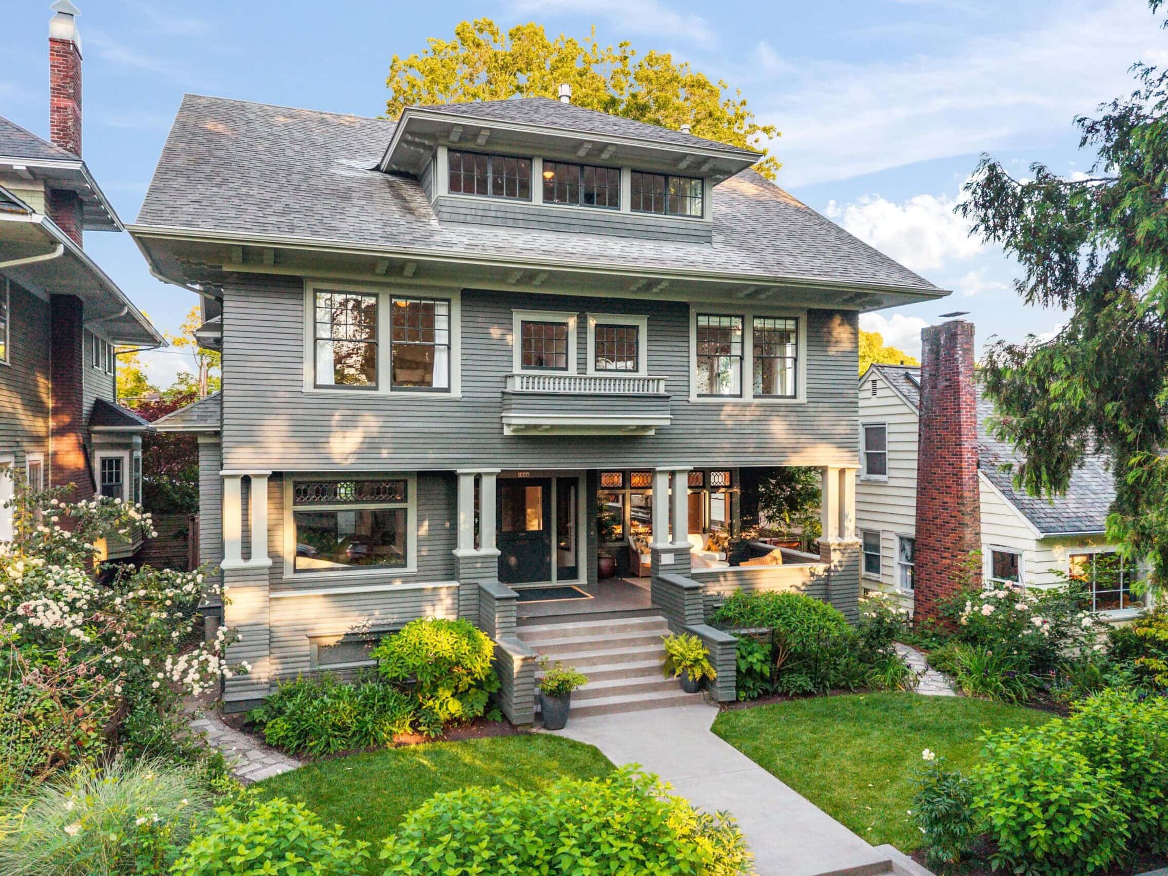A large, two-story gray craftsman-style house with a covered front porch, surrounded by lush green shrubs, plants, and trees, with a concrete walkway leading to the steps.