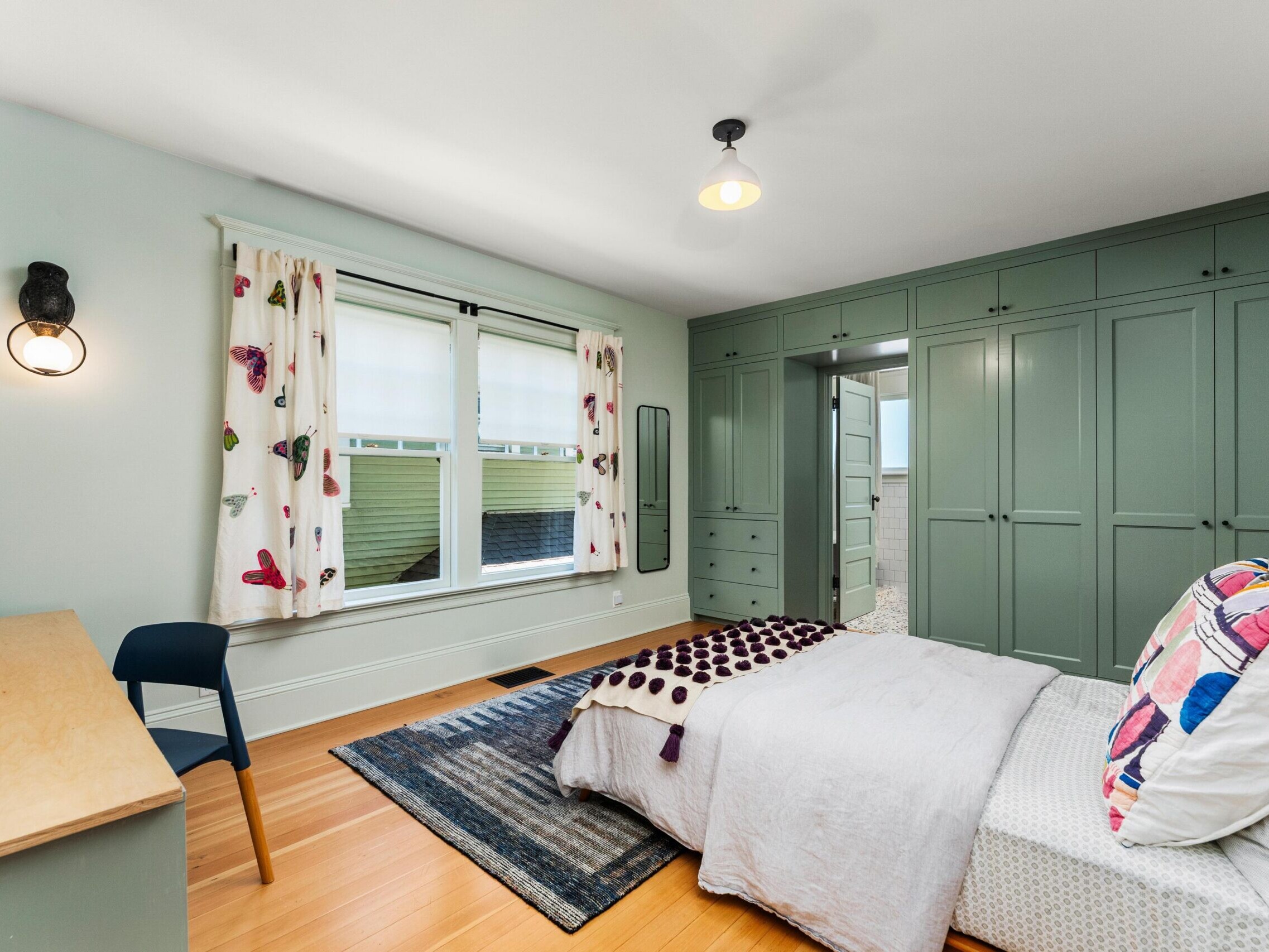 A bright bedroom with green built-in wardrobes, a bed with patterned pillows and blanket, a wooden desk with a black chair, butterfly curtains on a window, and a gray rug on a wood floor.