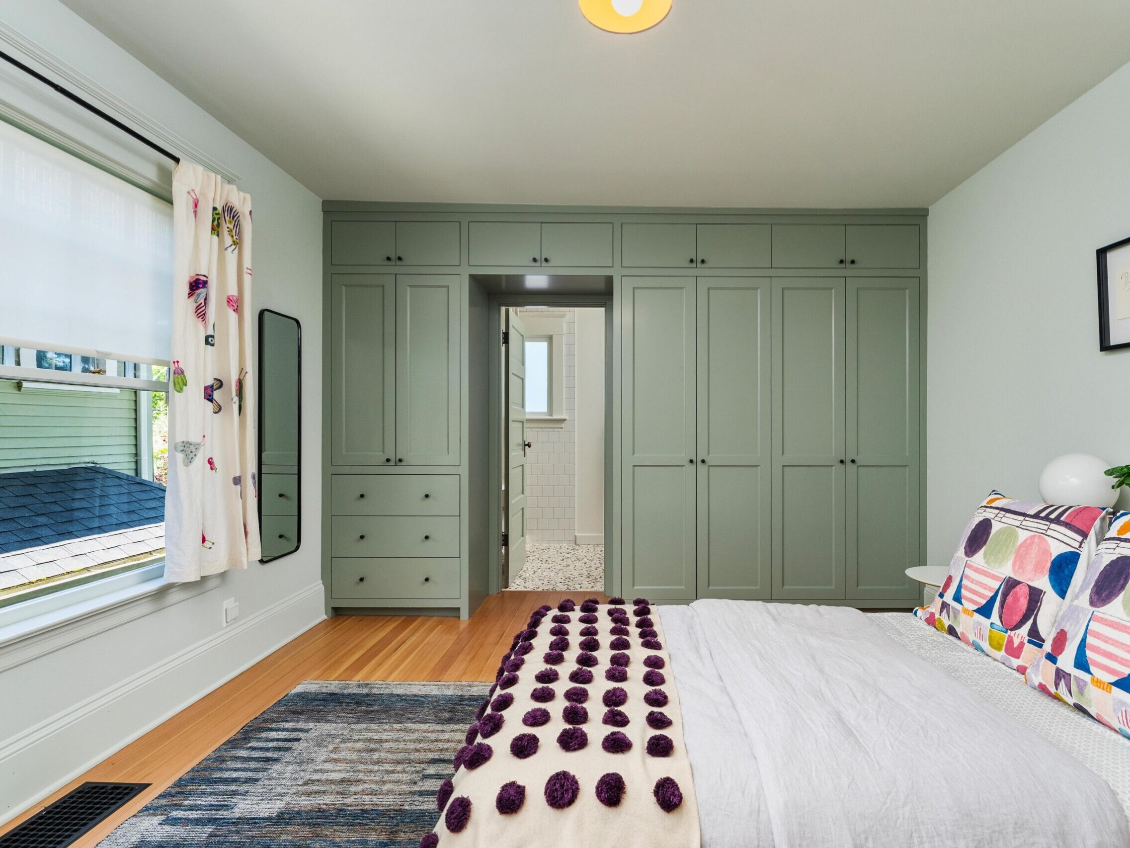 A bright bedroom with sage green built-in wardrobes, a bed with colorful geometric pillows and a pom-pom blanket, a window with butterfly curtains, a wall mirror, and an open doorway leading to a bathroom.