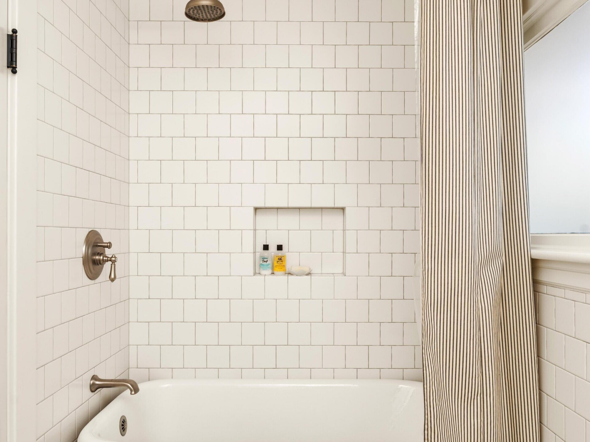 A bathroom with white tiled walls, a dark purple clawfoot bathtub, a striped shower curtain, and a rainfall showerhead. There are two small bottles in a recessed shelf and the floor is covered in colorful mosaic tiles.
