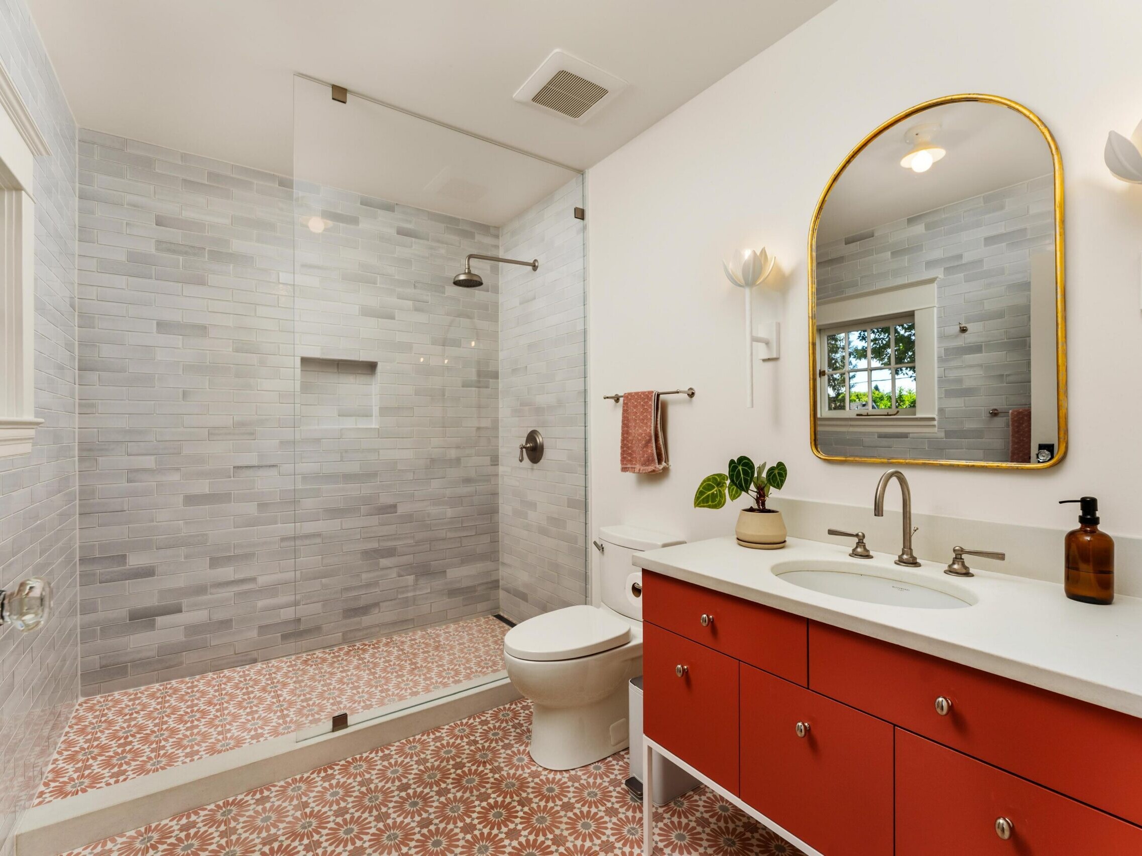 Modern bathroom with a glass-enclosed shower, white and gray tile walls, a toilet, and a vanity with a red cabinet, white countertop, oval mirror, and two wall sconces. Patterned floor tiles and a window provide natural light.