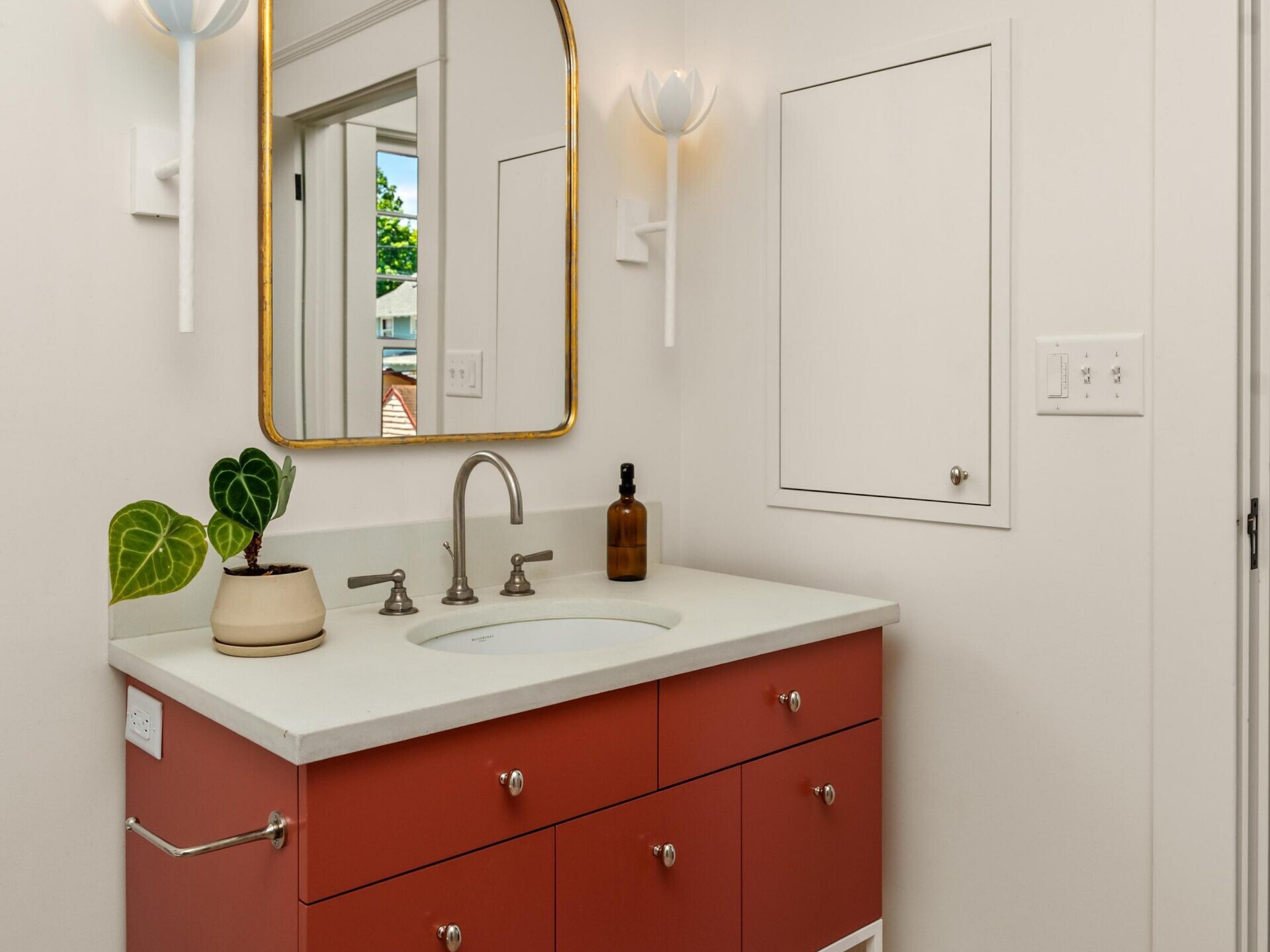 A modern bathroom features a red vanity with six drawers, a white countertop, and a silver faucet. Above it hangs an arched mirror with a gold frame, flanked by two white wall sconces. The floor has a red and white floral tile pattern.