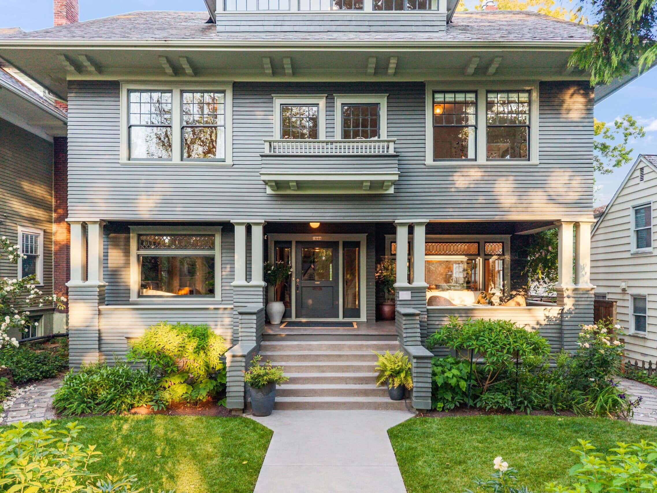 A large, two-story house with gray siding, a covered front porch, wide steps, and potted plants, surrounded by green shrubs and a manicured lawn.