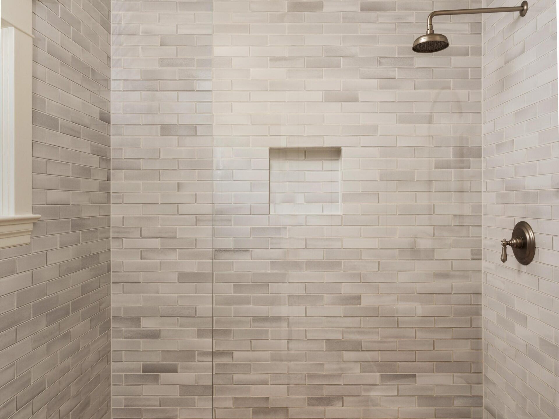 A modern walk-in shower with light gray subway tiles, a built-in shelf, a glass divider, a bronze showerhead, and a colorful red and white patterned tile floor.