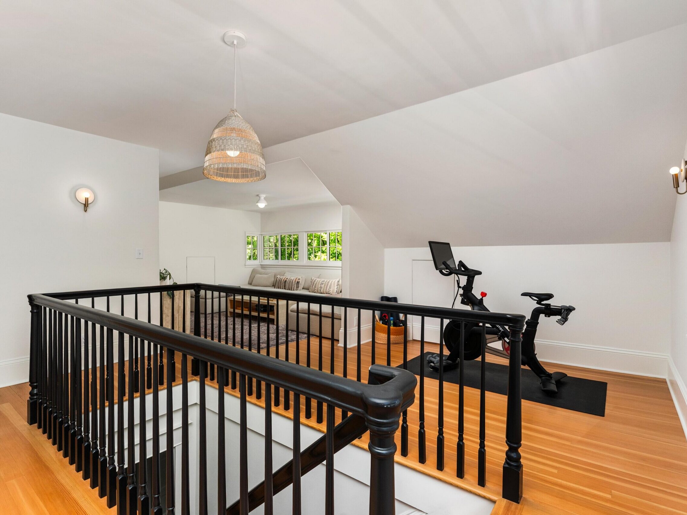 A bright upstairs loft with wood floors, a black exercise bike, a cozy seating area with a beige sofa, black railings around the staircase, and white walls with modern light fixtures.