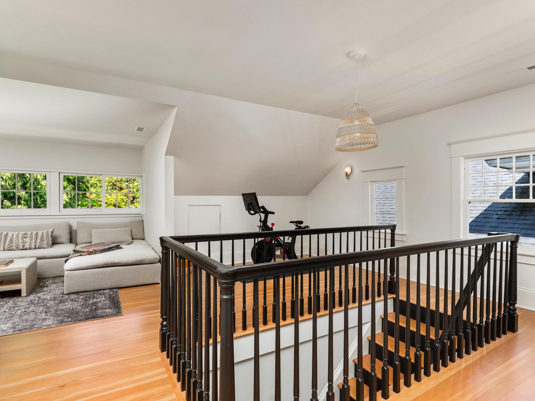 A bright upstairs landing with hardwood floors, a black staircase railing, a stationary bike, and a cozy sitting area with a sectional sofa and large windows letting in natural light.