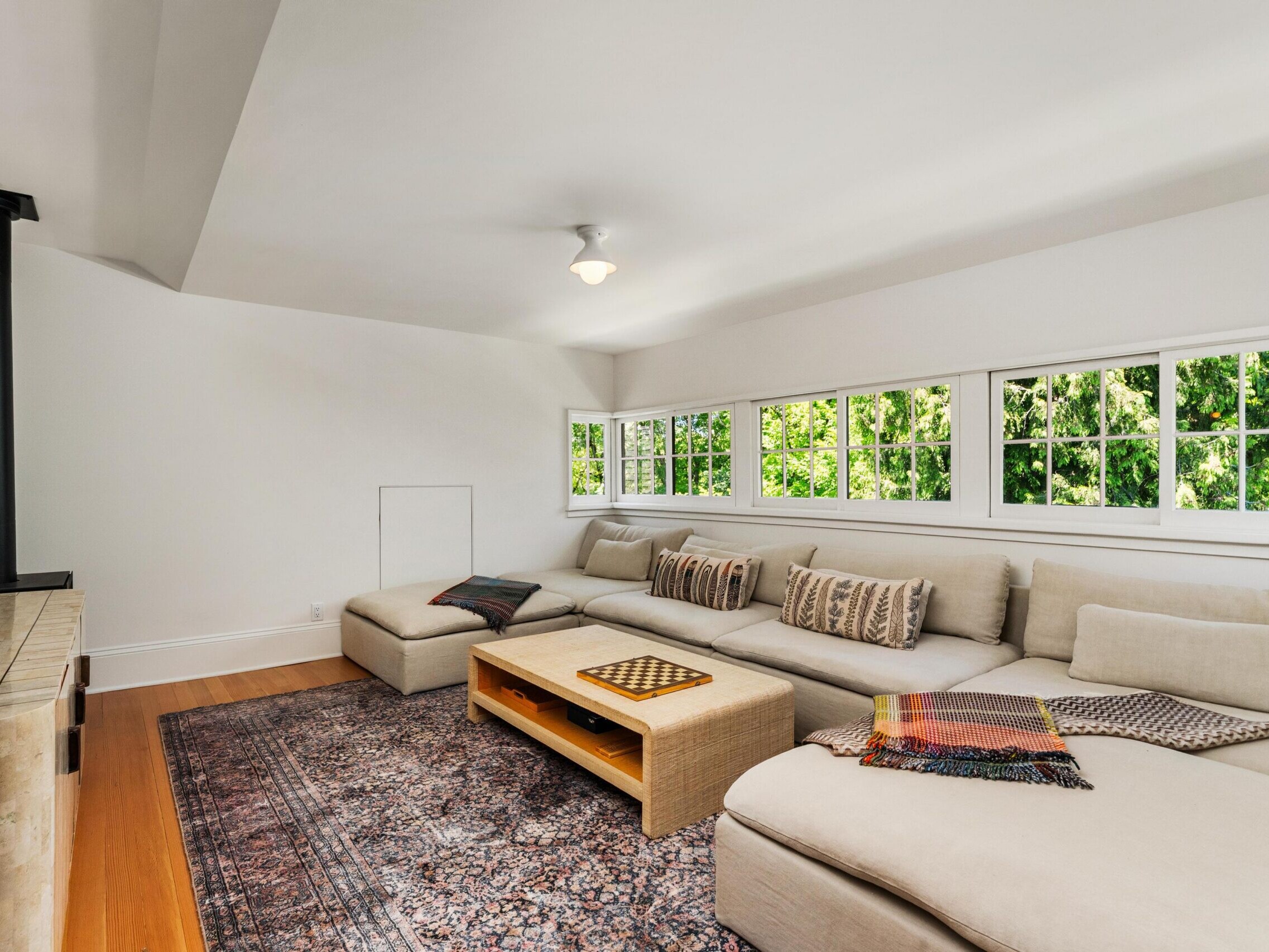 A spacious, bright living room with a large beige sectional sofa, patterned pillows, blankets, a wooden coffee table, a patterned rug, and multiple windows letting in natural light with a view of green trees outside.