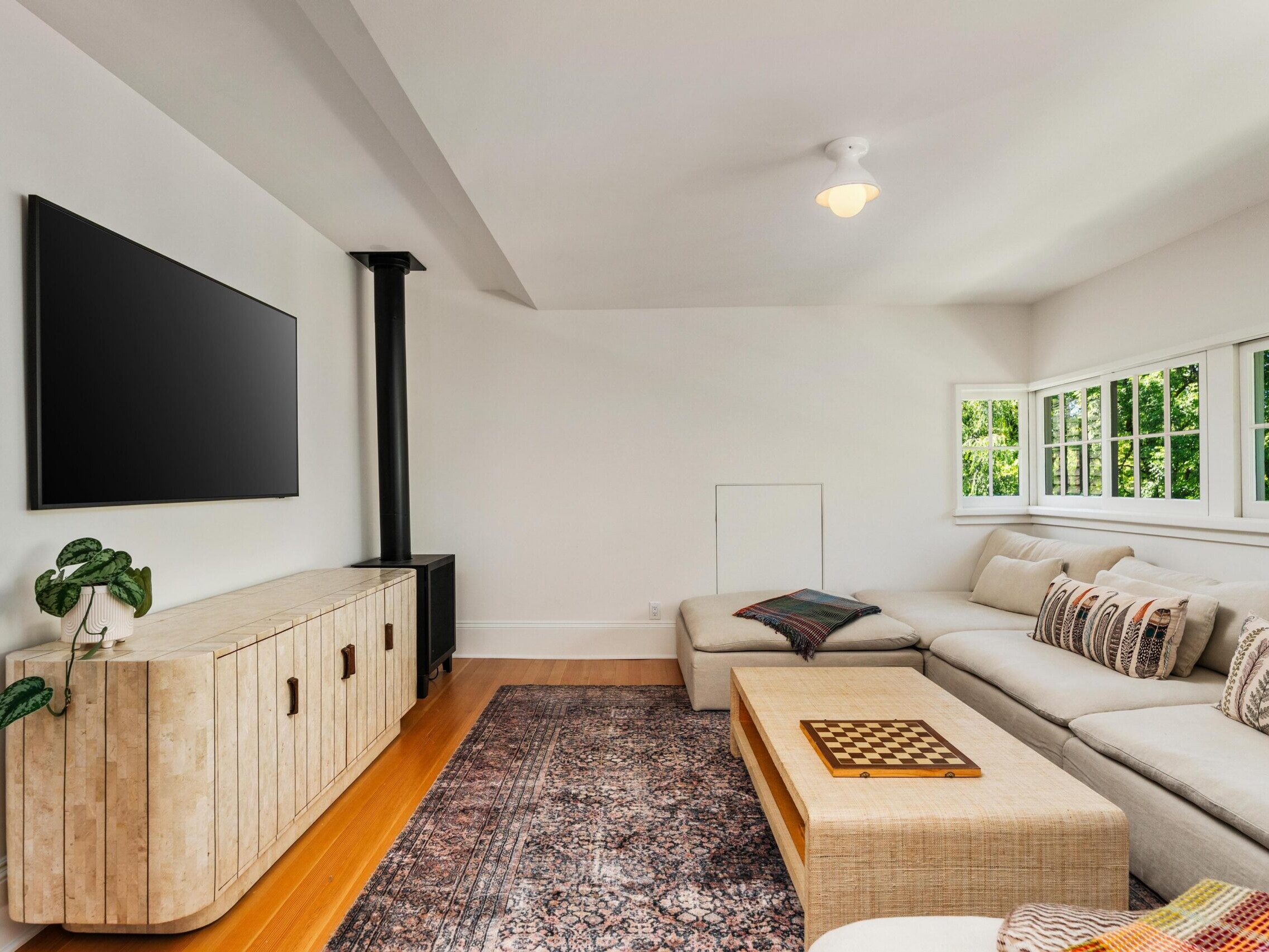 A bright, modern living room with a large beige sectional sofa, patterned pillows, a wooden coffee table with a chessboard, a textured console under a wall-mounted TV, and windows letting in natural light.