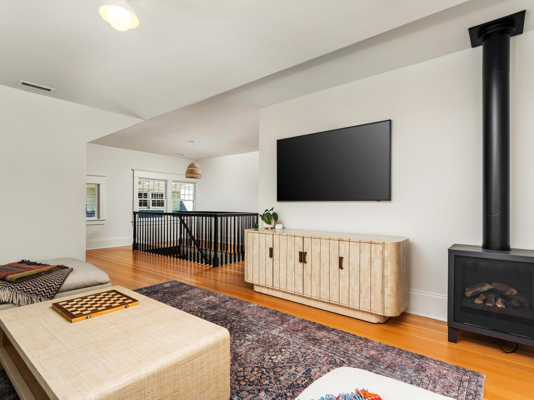 A modern living room with a large wall-mounted TV, a light wood cabinet, a black wood-burning stove, a sectional sofa, a patterned rug, and railing by a staircase. Natural light comes in through nearby windows.