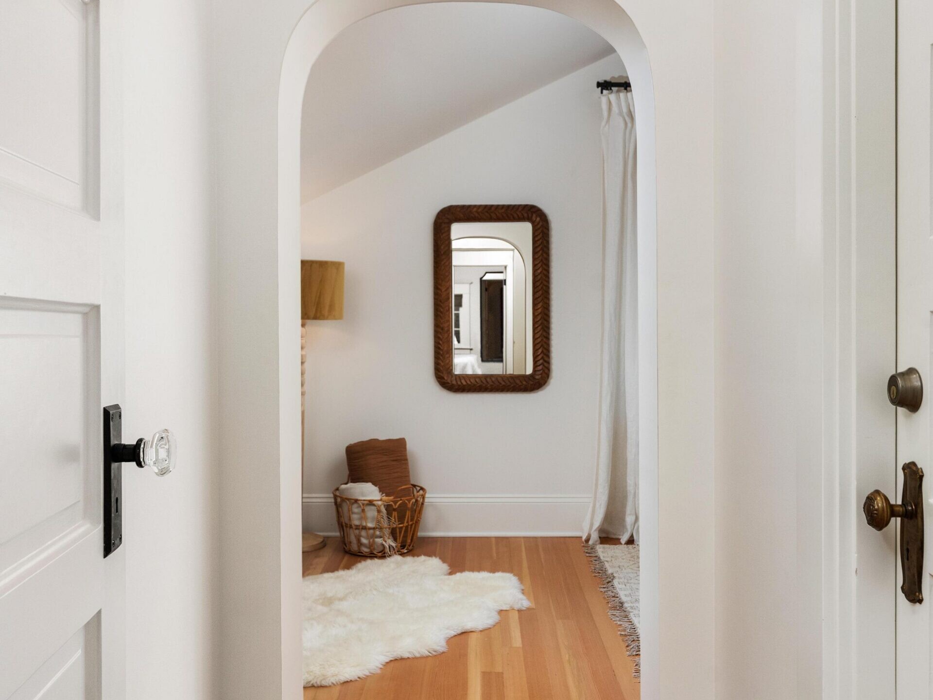 A hallway with light wood flooring and white walls leads through an arched doorway to a cozy room with a sheepskin rug, basket, lamp, and a framed mirror reflecting the hallway.