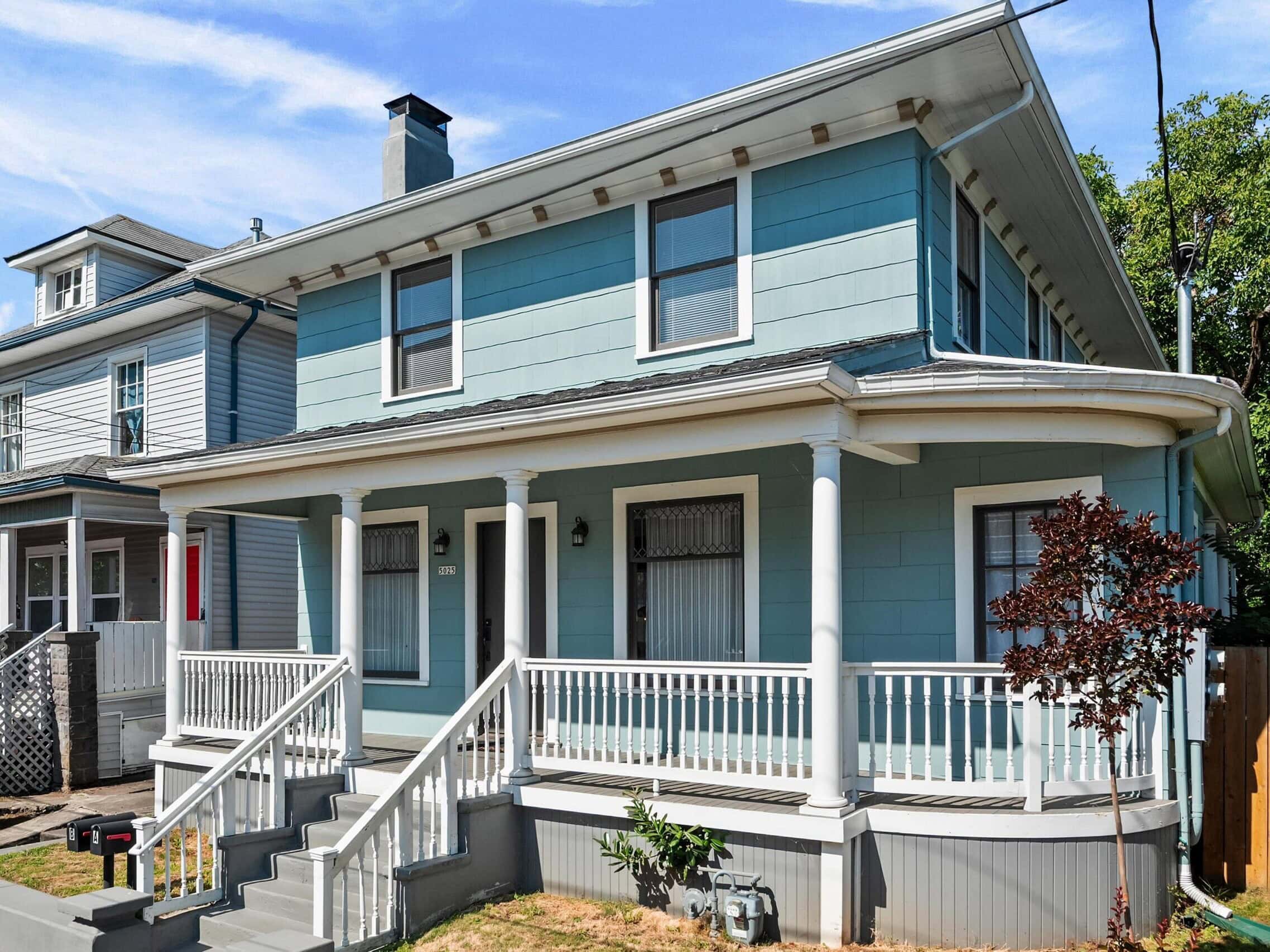 A two-story blue house with white trim, a covered front porch, gray steps, and a small front yard. There are trees and another house visible in the background.