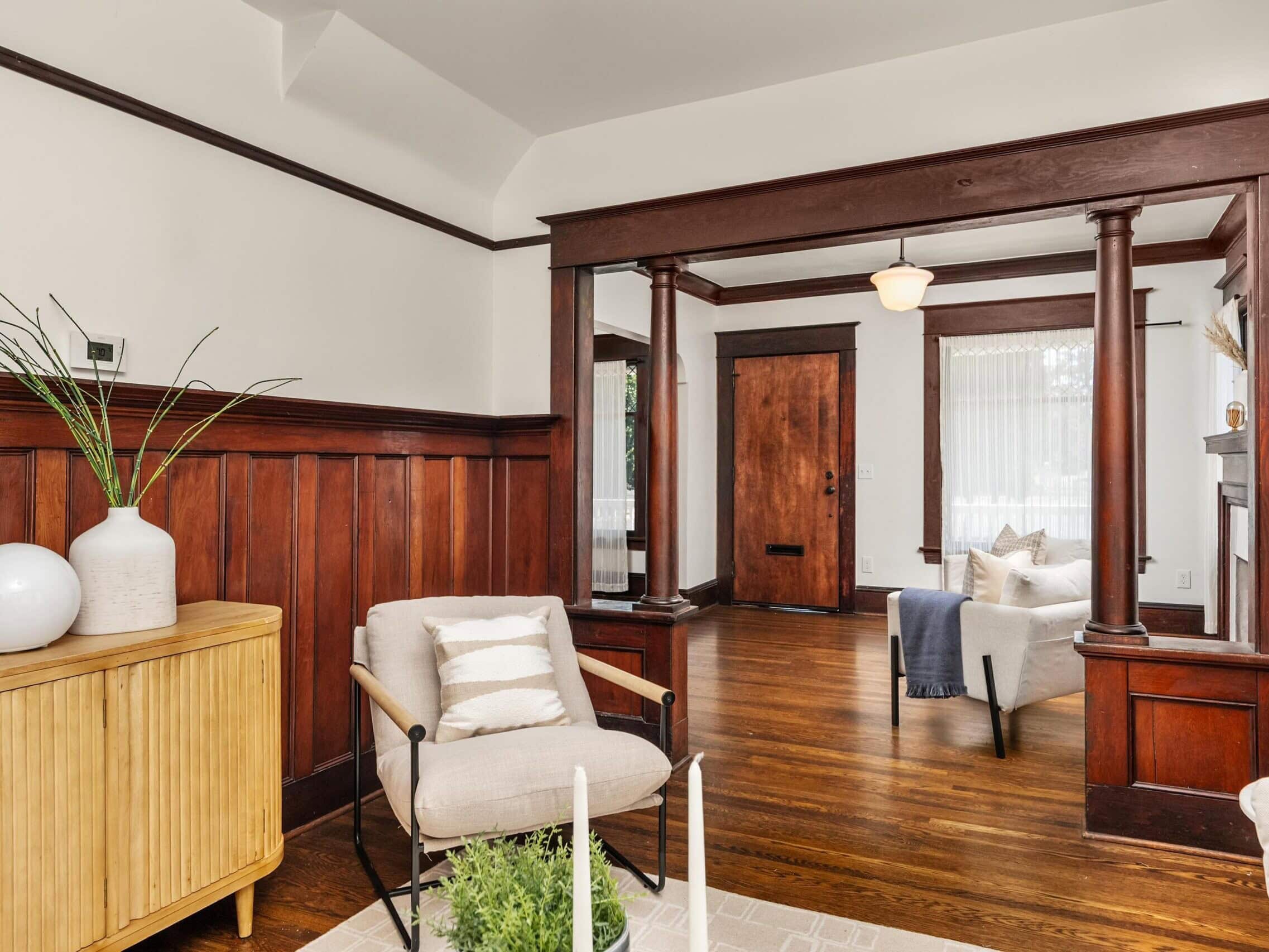 Living room with wood paneling, hardwood floors, an armchair, sideboard with decor, and a view of a wooden front door and another seating area in the background.