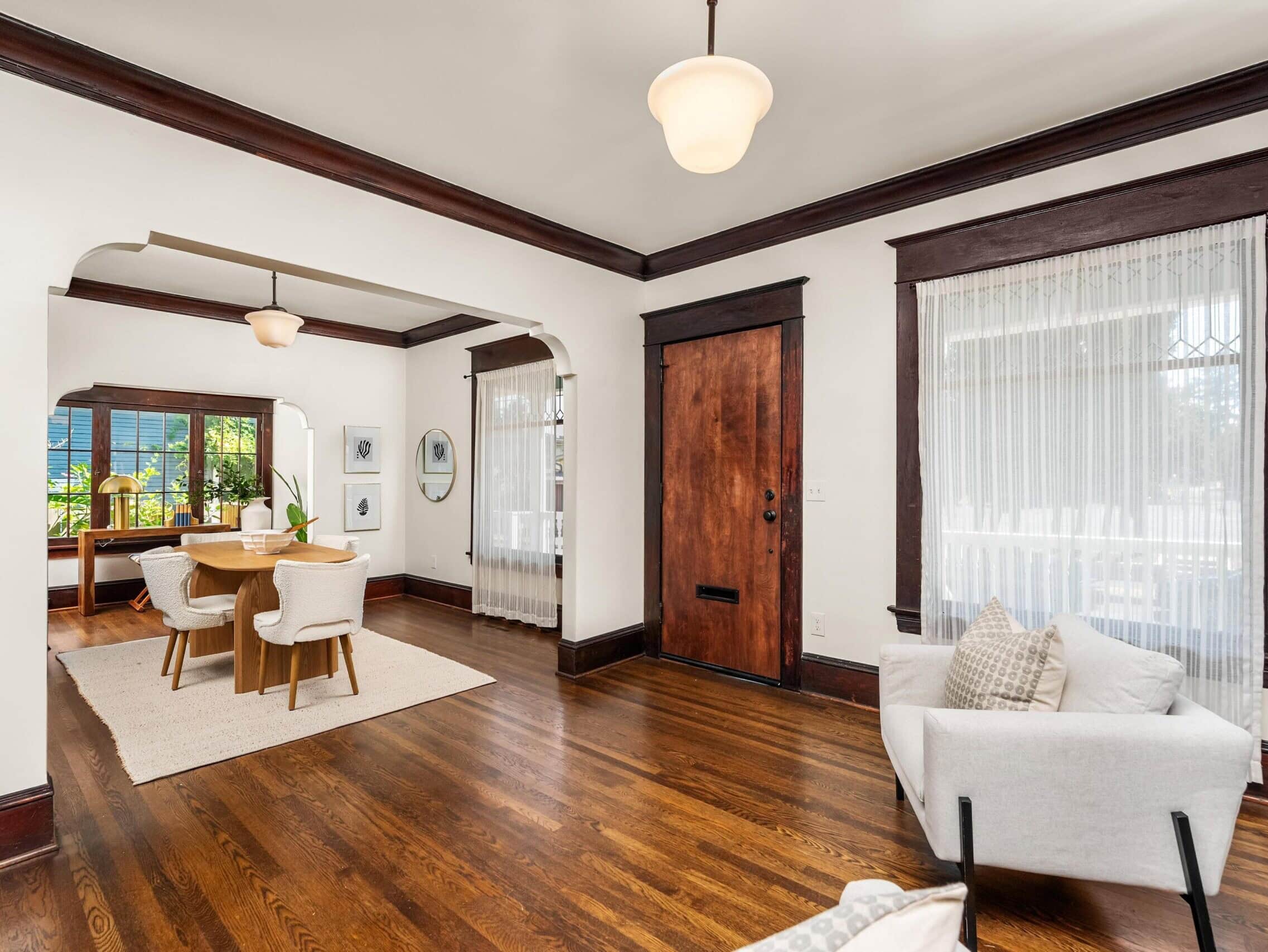 Bright living and dining area with hardwood floors, white walls, dark wood trim, and large windows with sheer curtains. Features a round dining table with chairs and cozy armchairs.