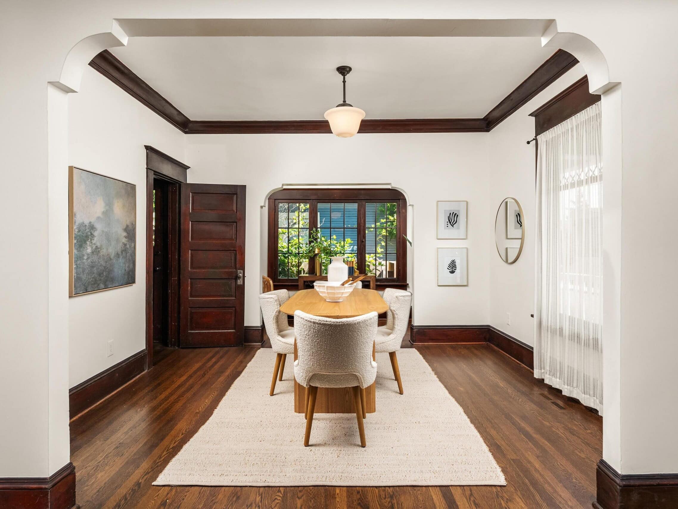 A bright dining room with a wooden table, six white chairs, artwork on white walls, dark wood trim, sheer curtains, and a window letting in natural light.
