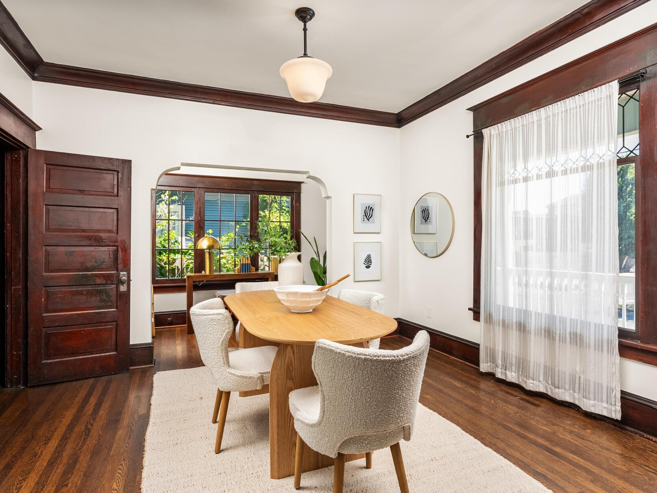 A bright dining room with a wooden table, four white textured chairs, hardwood floors, sheer white curtains, dark wood trim, and wall art. A sideboard with plants is visible in the background through an open doorway.