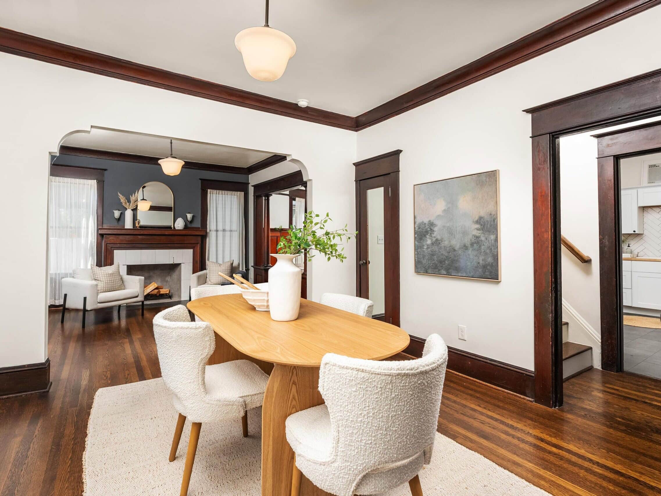 A modern dining room with a wooden table and white chairs, hardwood floors, white walls with dark trim, and a view into a living room with a fireplace and a kitchen with white cabinets.