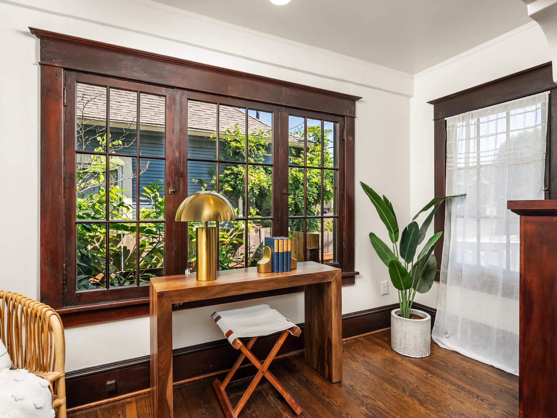 A wooden desk with a gold lamp, books, and decor sits by large windows with dark trim. A potted plant and a woven chair are nearby, and sunlight streams in, brightening the cozy space.
