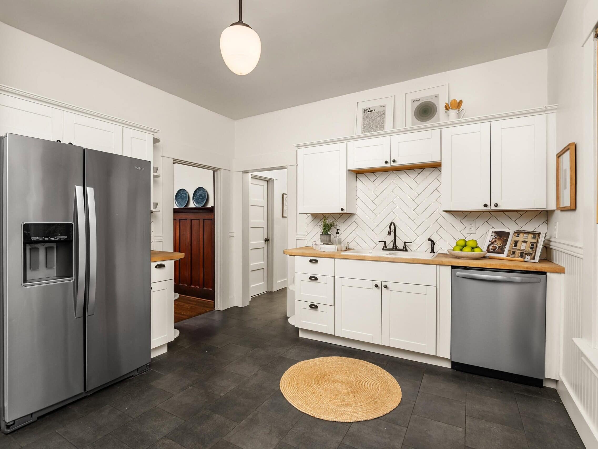 Modern kitchen with white cabinets, stainless steel appliances, a round woven rug, wooden countertops, and chevron tile backsplash. A window lets in natural light, and shelves hold decor items and dishes.