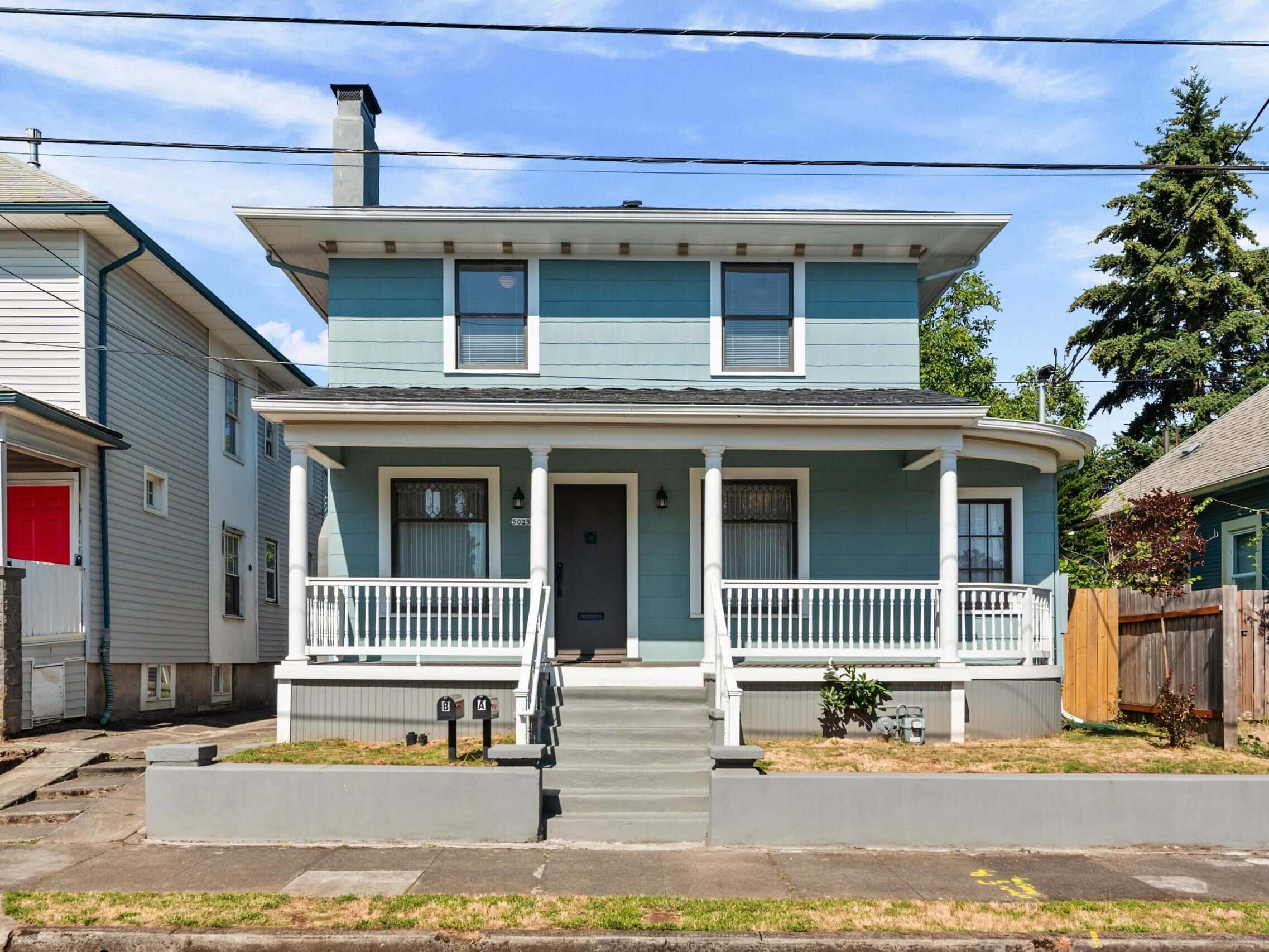 A two-story blue house with white trim, a covered front porch, steps, railing, and a small lawn. The house is between two other homes on a residential street under a partly cloudy sky.