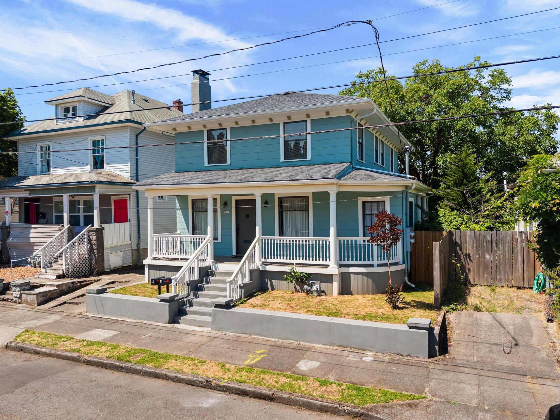 Two-story blue house with white trim, a covered front porch, and steps leading to the entrance. The house has a fenced backyard and is situated on a residential street with neighboring homes.