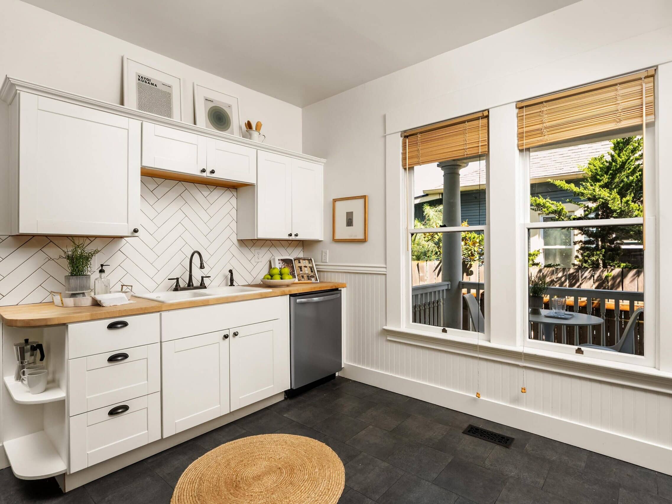 Bright kitchen with white cabinets, wood countertops, a small round rug, window with bamboo blinds, and a view of a porch and greenery outside. Minimalist decor and modern, clean design.