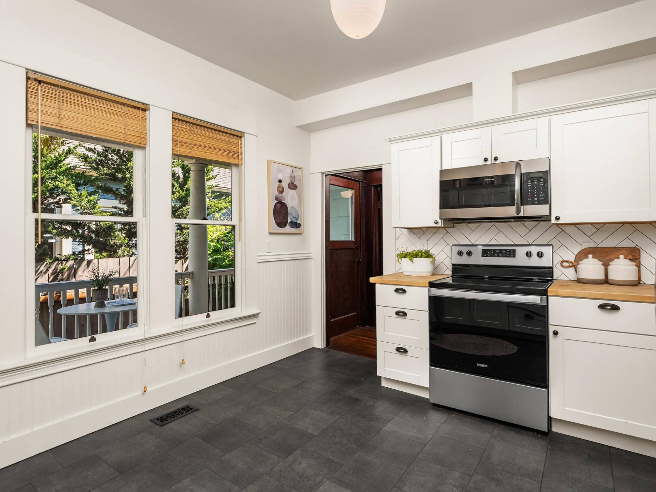 Bright kitchen with white cabinets, black appliances, wood countertops, large windows, and a view of a patio table outside. Theres a wall art piece and three ceramic canisters on the counter.