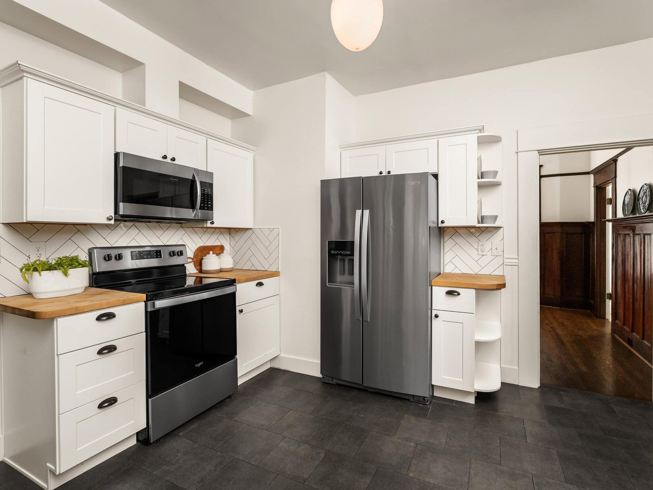 Modern kitchen with white cabinets, wood countertops, black stove, built-in microwave, stainless steel refrigerator, potted plant, and geometric tile backsplash. Doorway leads to a hallway with wood paneling.
