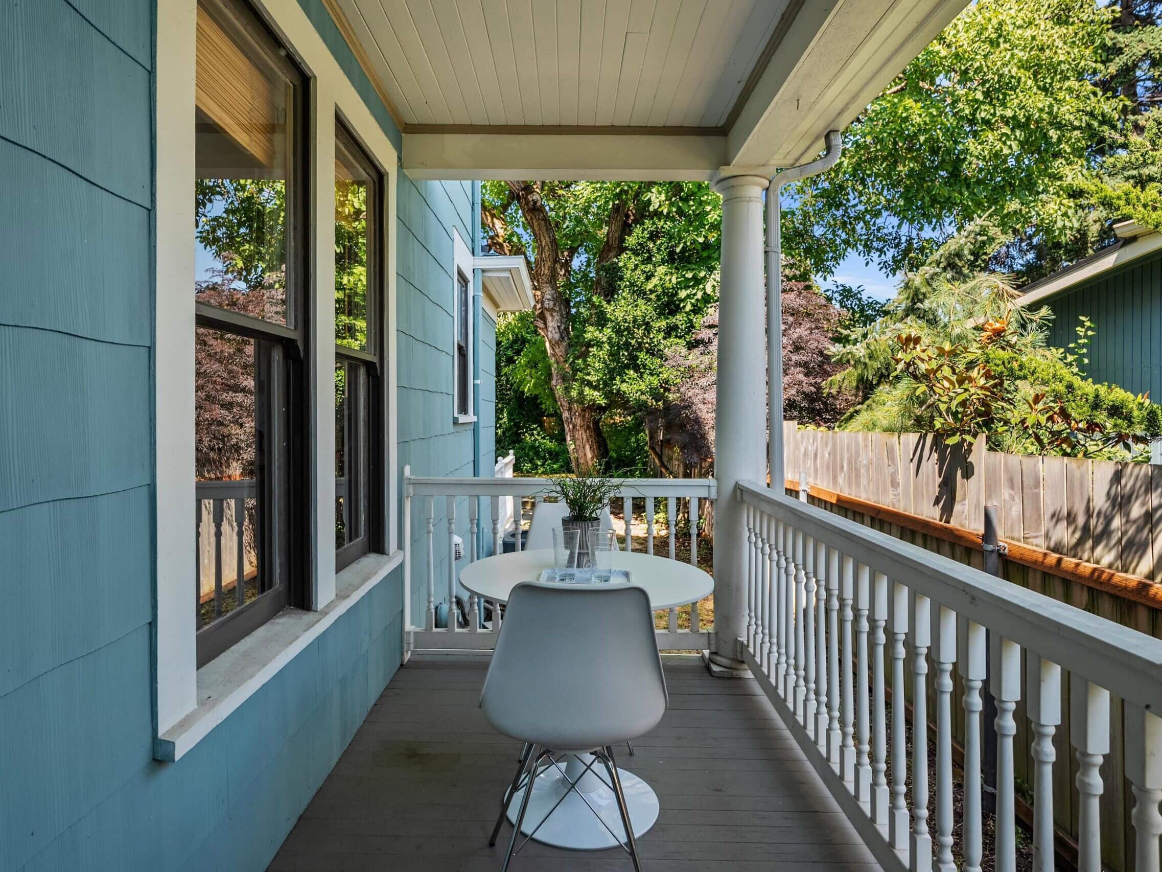 Small porch with blue walls, white railing, a round white table, and a single modern chair. Trees and greenery are visible beyond the railing, providing a peaceful outdoor setting.